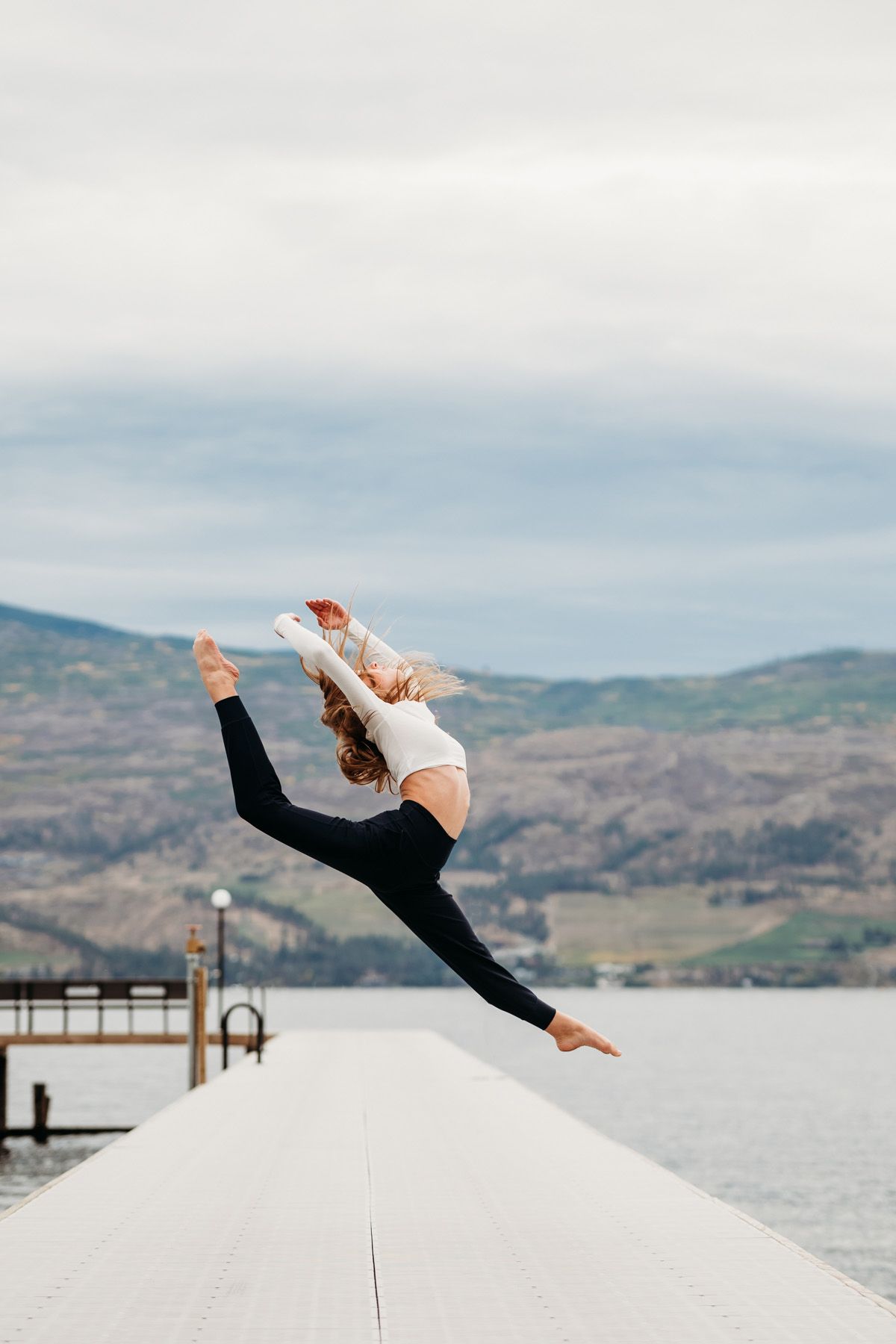 Dancer performs a dramatic split leap against a scenic mountain lake backdrop on a white platform.