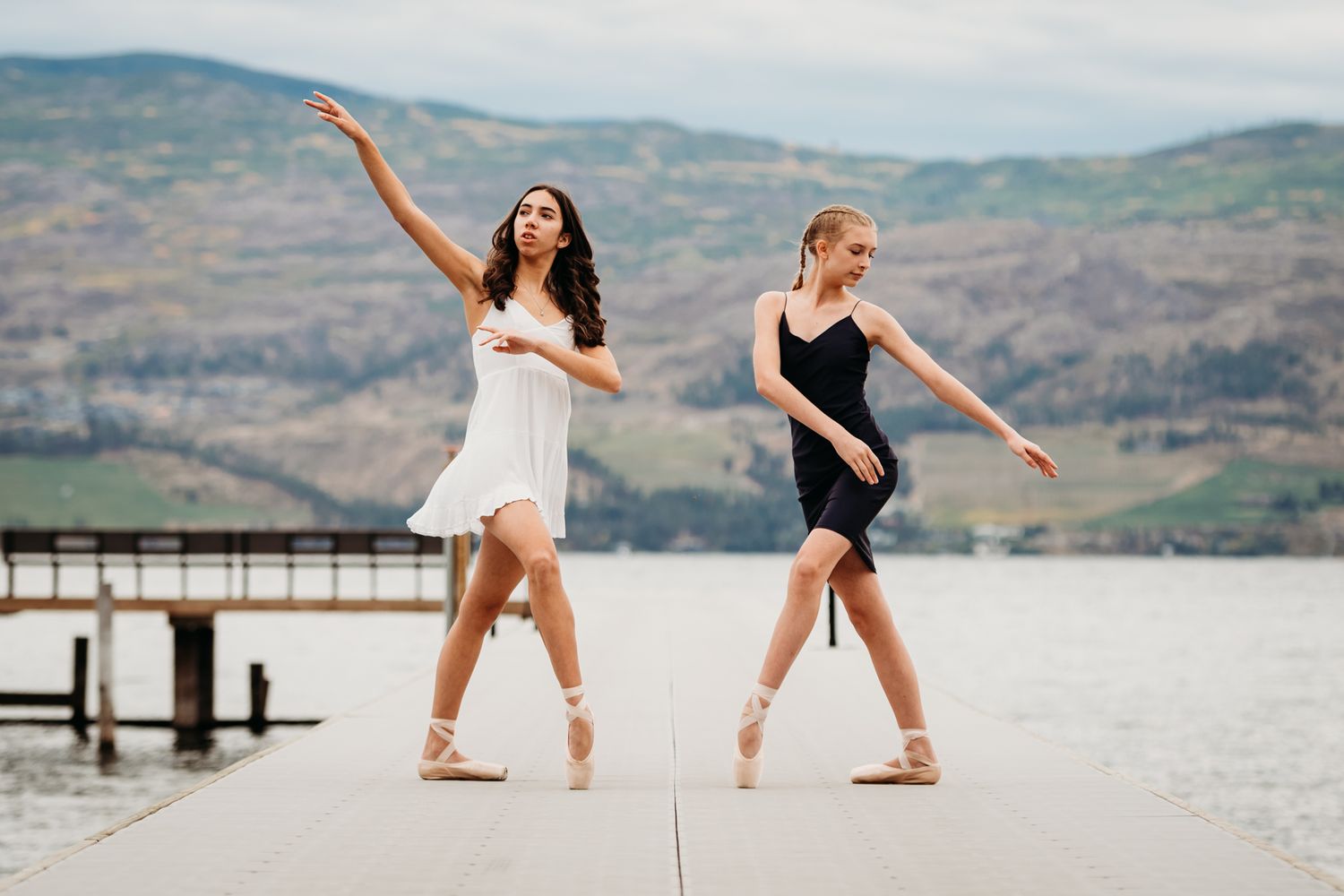 Two dancers in white and black dresses perform graceful ballet moves on a lakeside dock with mountains beyond.
