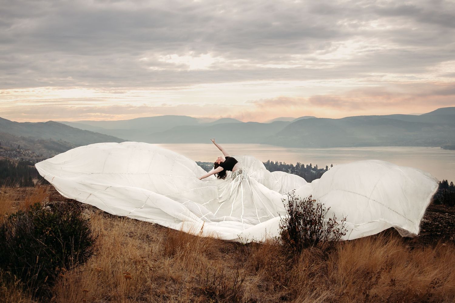 A paraglider soars over a scenic mountain lake with brown grass in the foreground under cloudy skies at sunset.