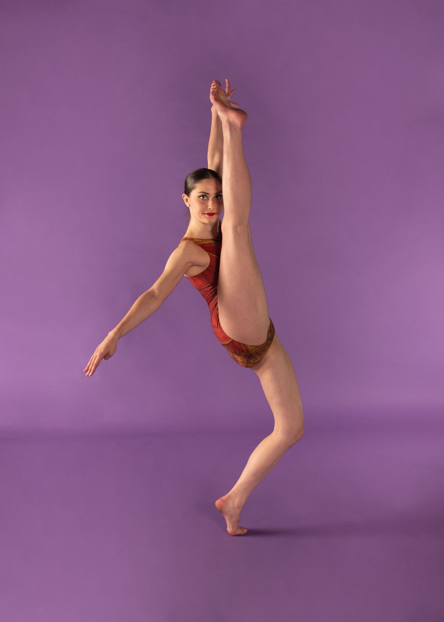 Dancer in red leotard performs graceful ballet pose against purple background, demonstrating flexibility and strength.