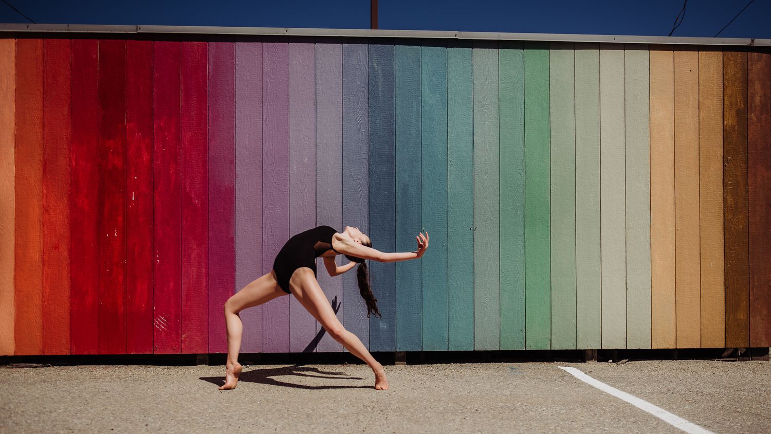Dancer in black leotard poses gracefully against vibrant rainbow-striped wall backdrop.