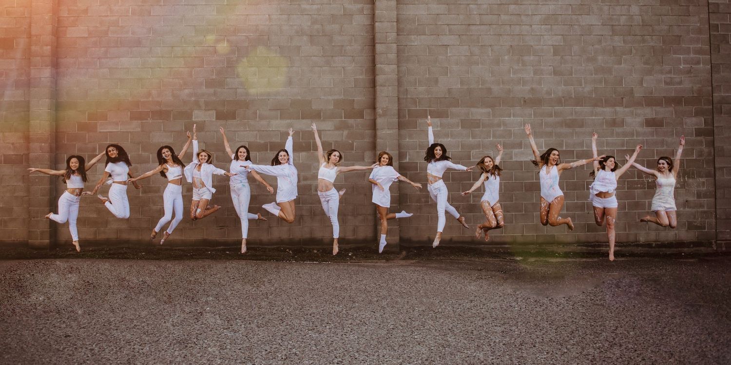 A group of ballet dancers in white dresses leaping gracefully in unison against a wall during sunset.