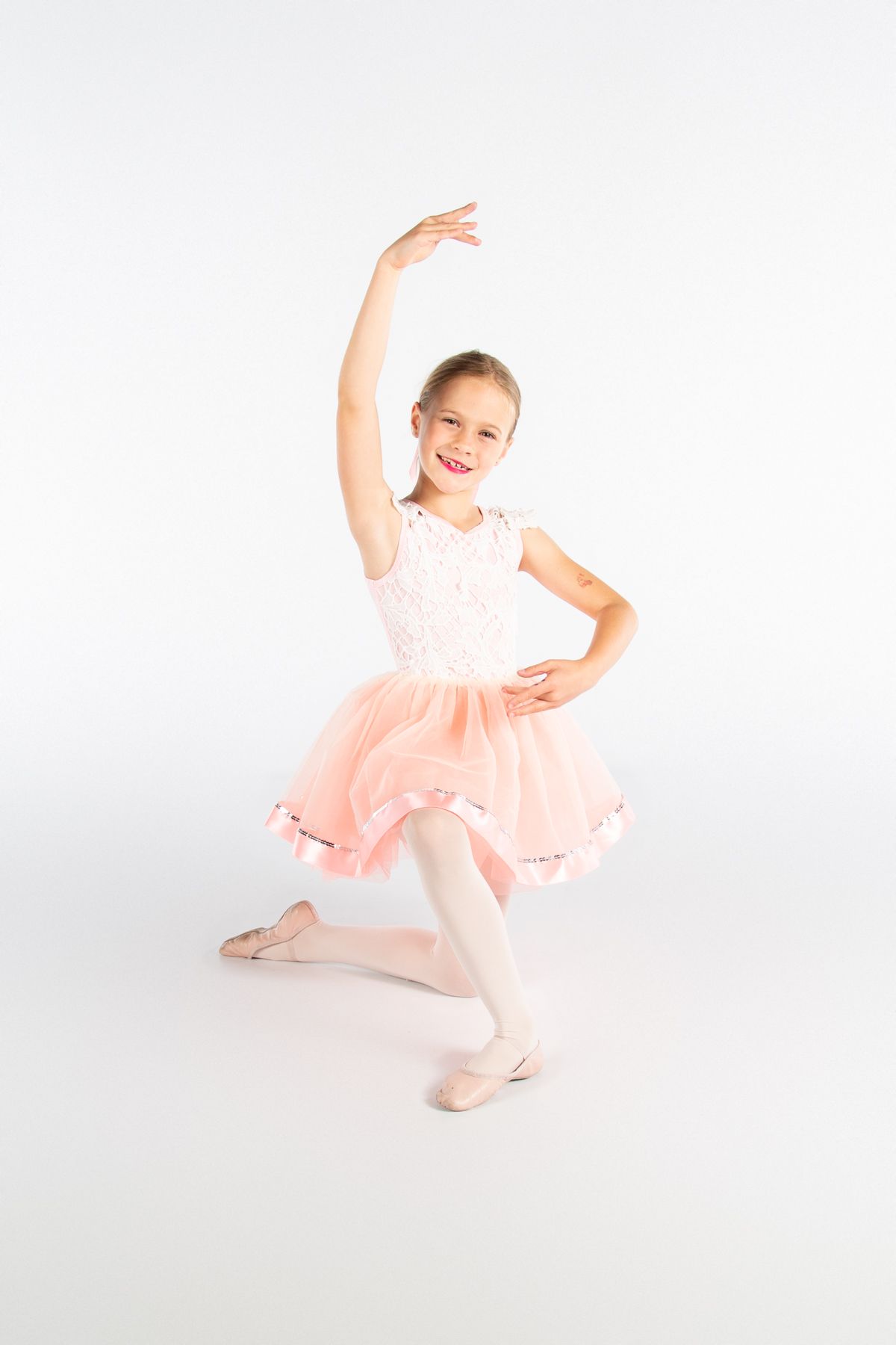 Young ballet dancer in pink tutu and white tights strikes graceful pose with uplifted arm on white background.