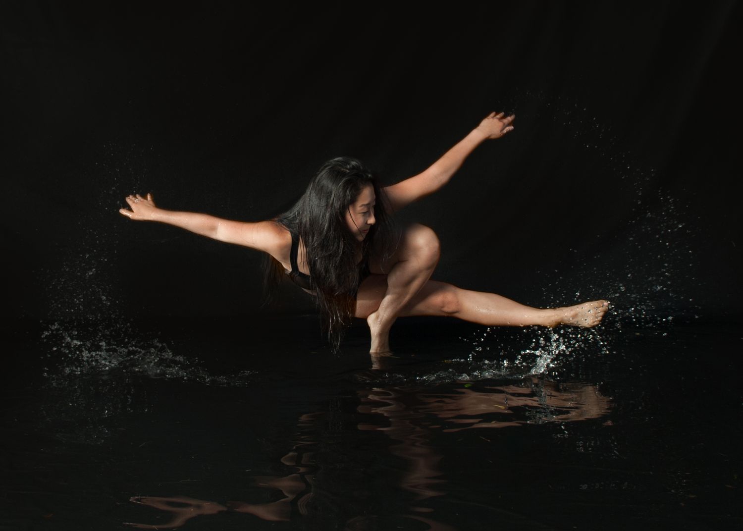 A dancer performs a dramatic pose in dark water creating splashes against a black background.