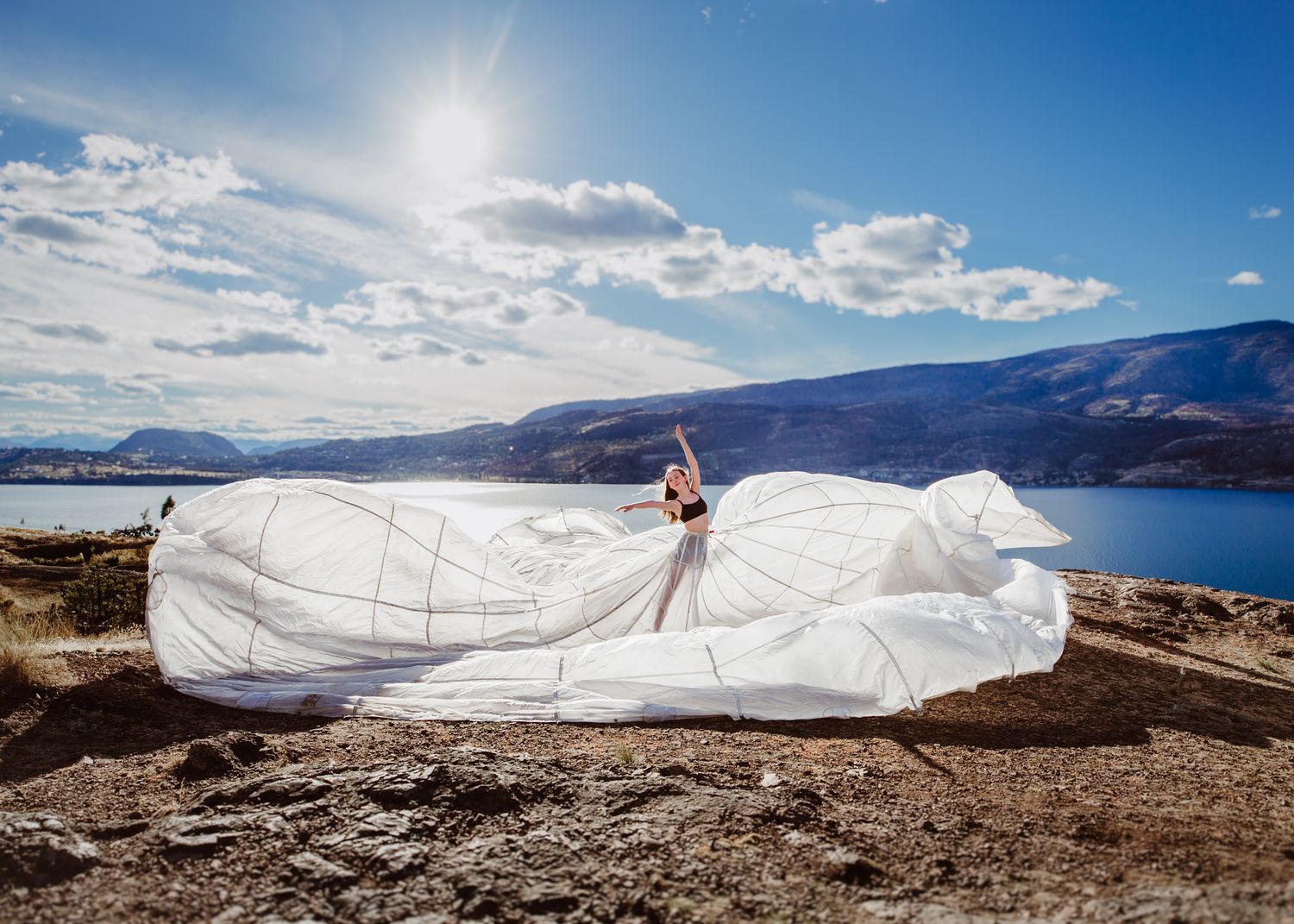 Flowing white dress billows dramatically against scenic mountain lake backdrop on sunny day with blue skies.
