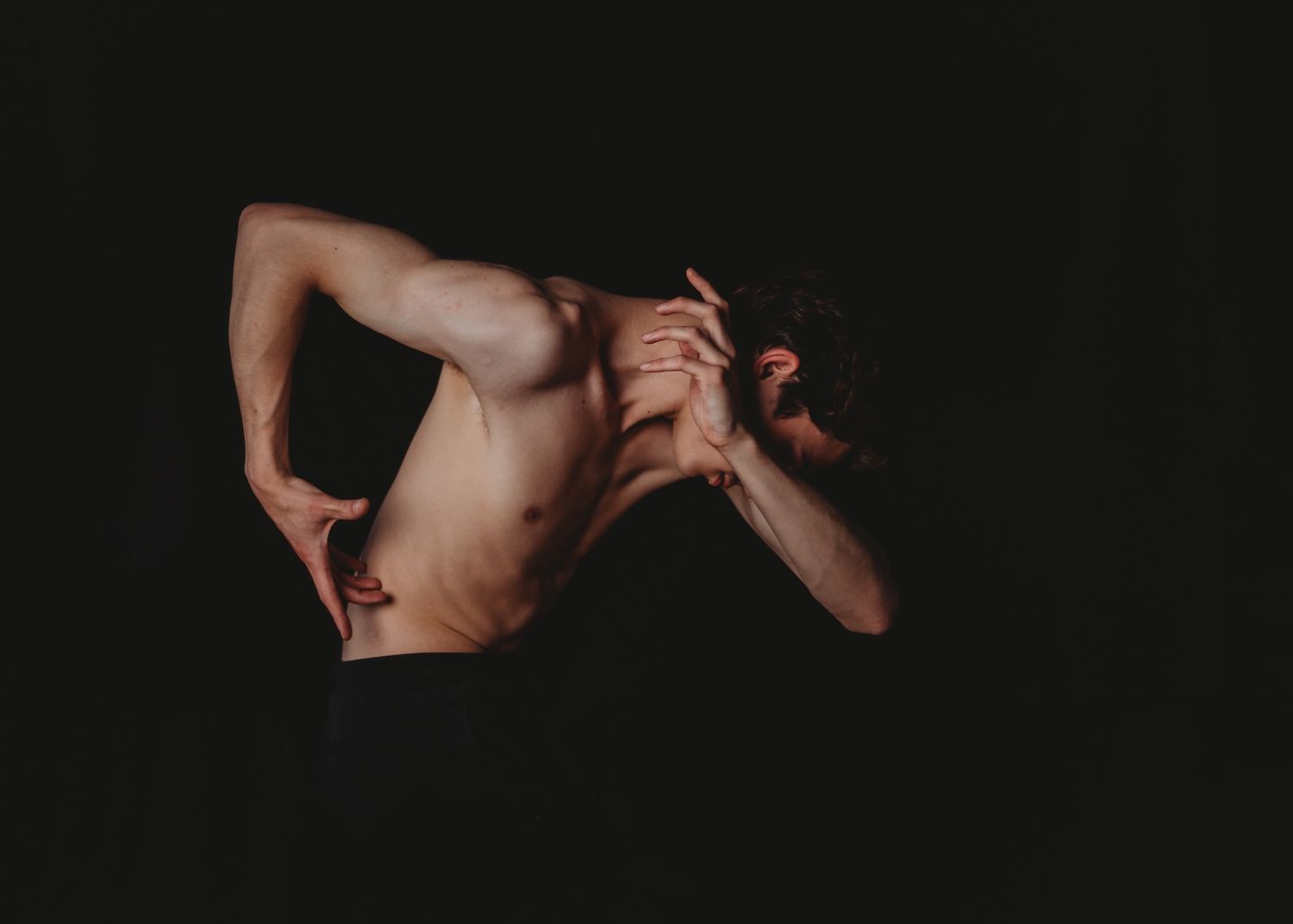 Artistic studio portrait of a shirtless torso and arm reaching against dark background in dramatic lighting.
