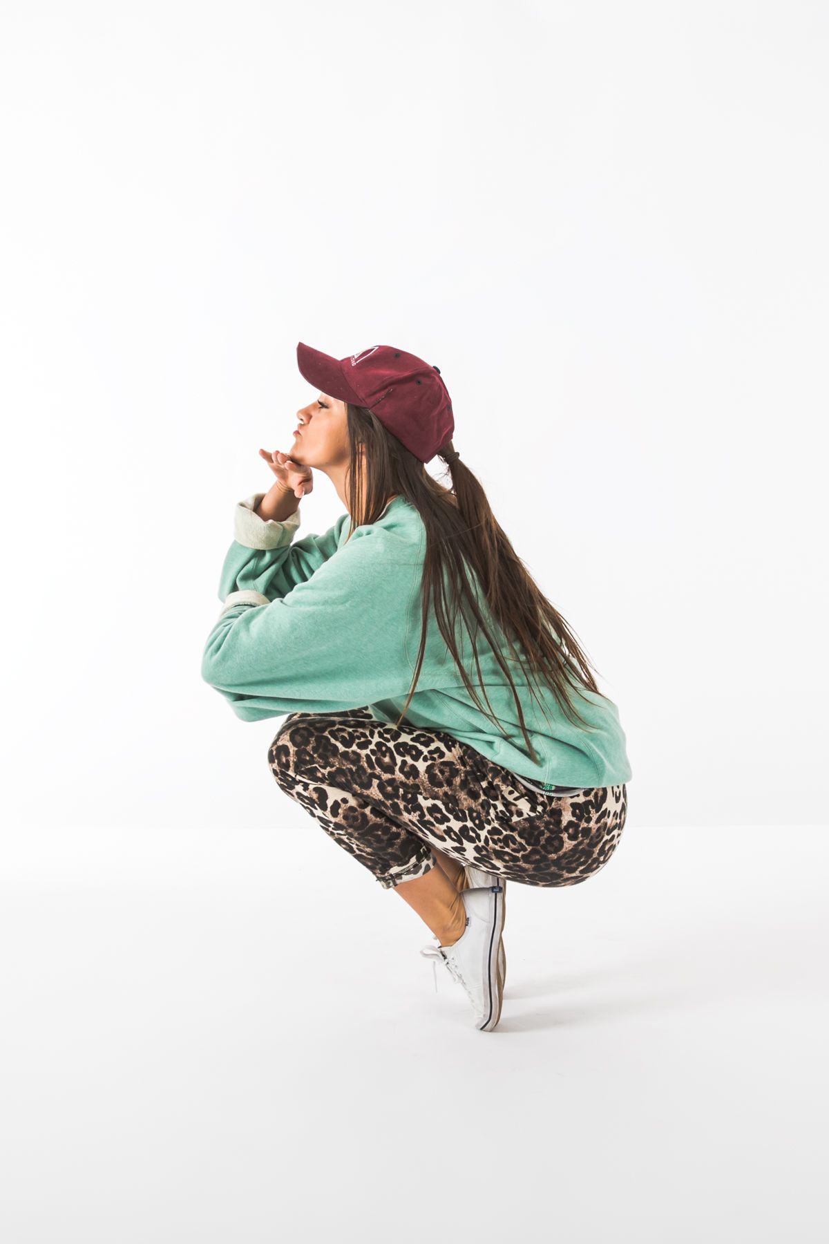 Dancer in mint sweater and leopard print pants crouches in urban style pose wearing burgundy cap.