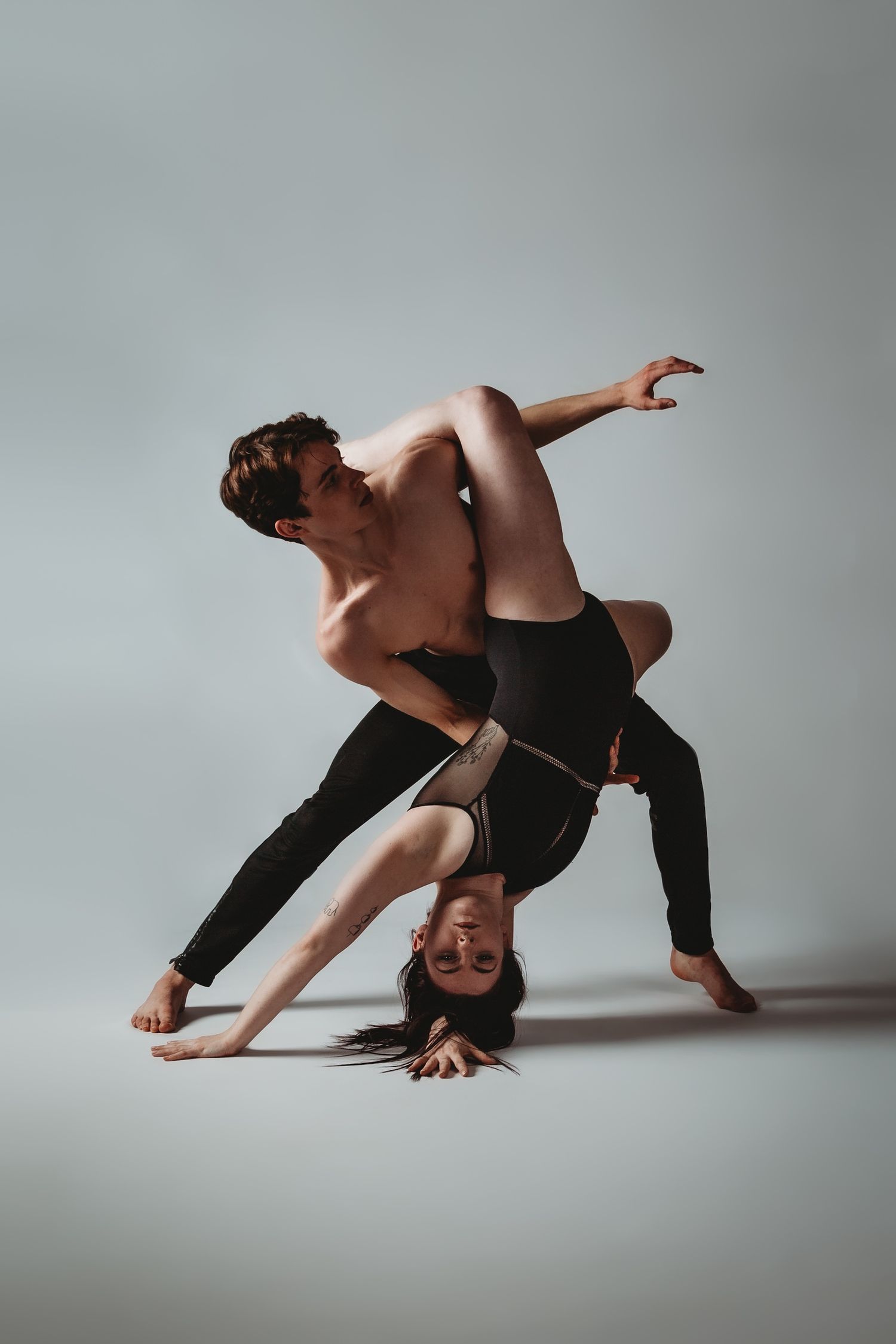 Two dancers in black performing a contemporary dance lift pose against a gray background in dramatic lighting.