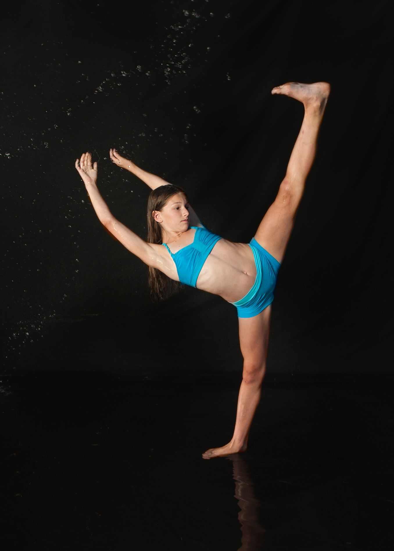 Dancer in bright blue athletic wear performs a high standing split against dark background with dramatic lighting.
