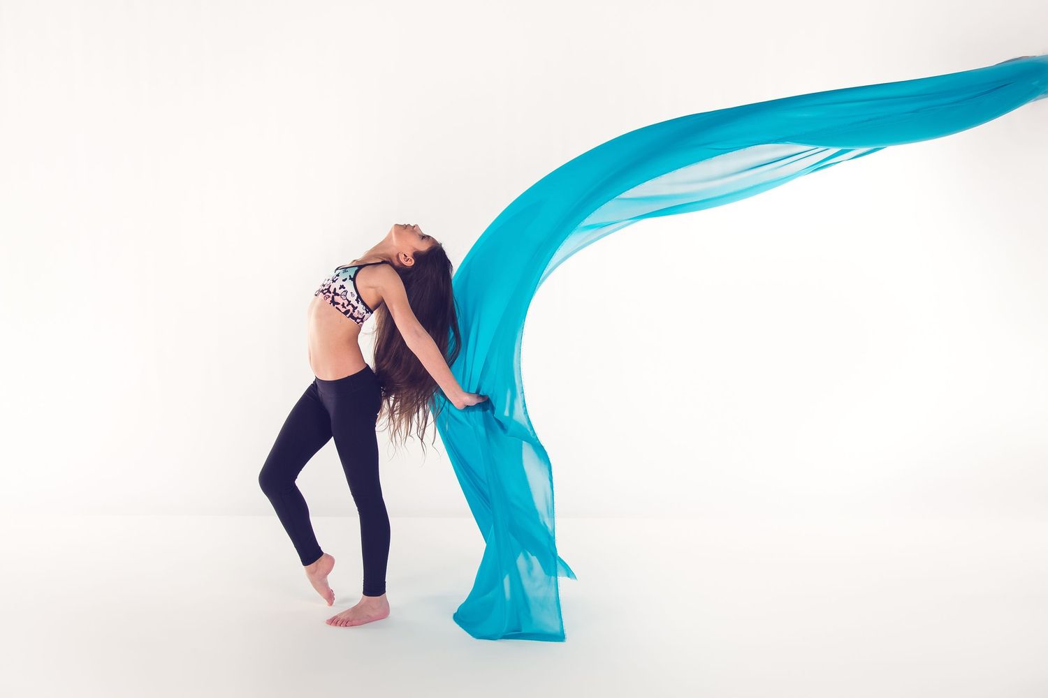 Dancer in dark leggings performs expressive movement with flowing turquoise fabric against white background.