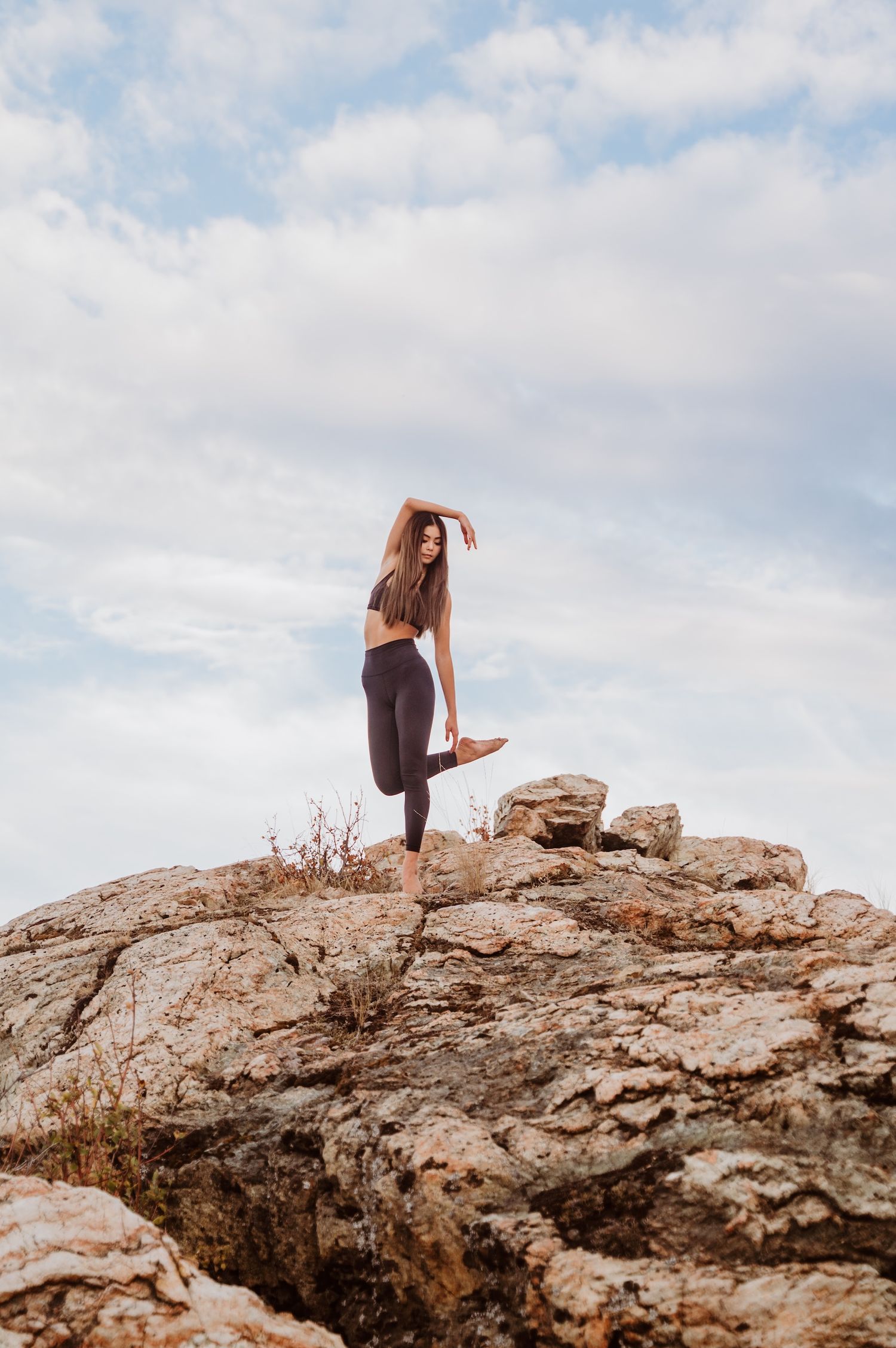 A dancer strikes a graceful pose on a rocky cliff edge against a cloudy sky.