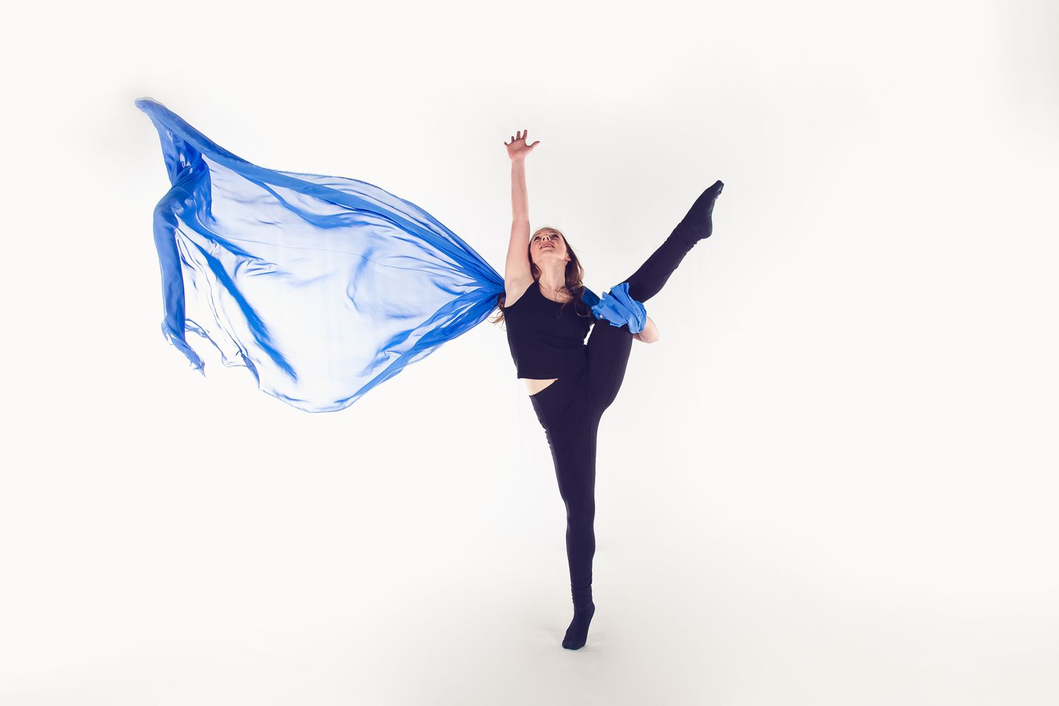 A dancer poses gracefully with a flowing blue fabric while performing an arabesque position against a white background.