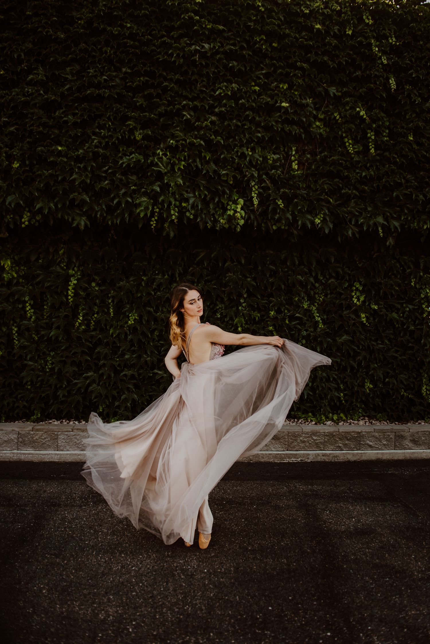 A flowing blush gown swirls gracefully as someone dances against a backdrop of dark hedges in moody lighting.