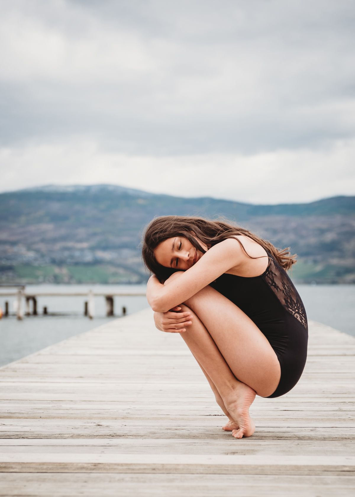 A person in a black outfit crouches on a wooden dock overlooking a serene lake with mountains in the background.