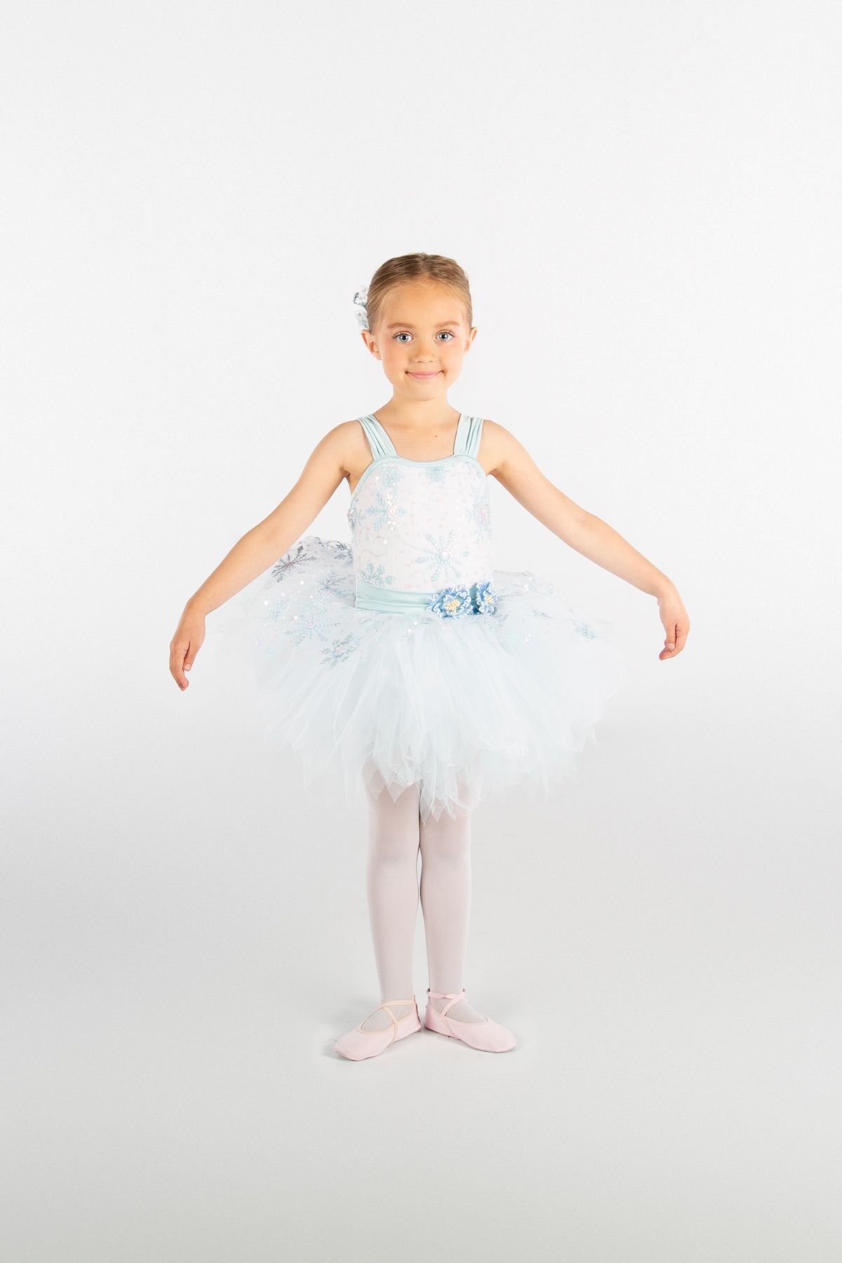 Young ballet dancer wearing light blue tutu and white leotard poses in first position against white backdrop.