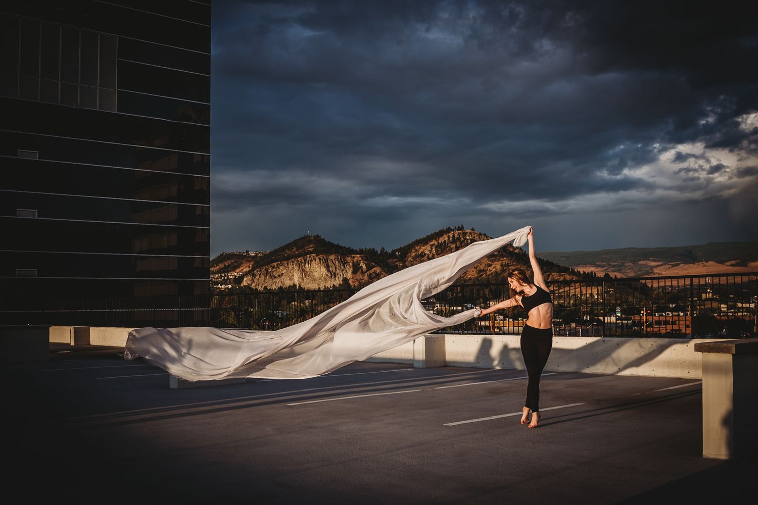 Dancer in motion on a rooftop creating a dramatic silhouette against stormy clouds with a flowing white fabric backdrop.