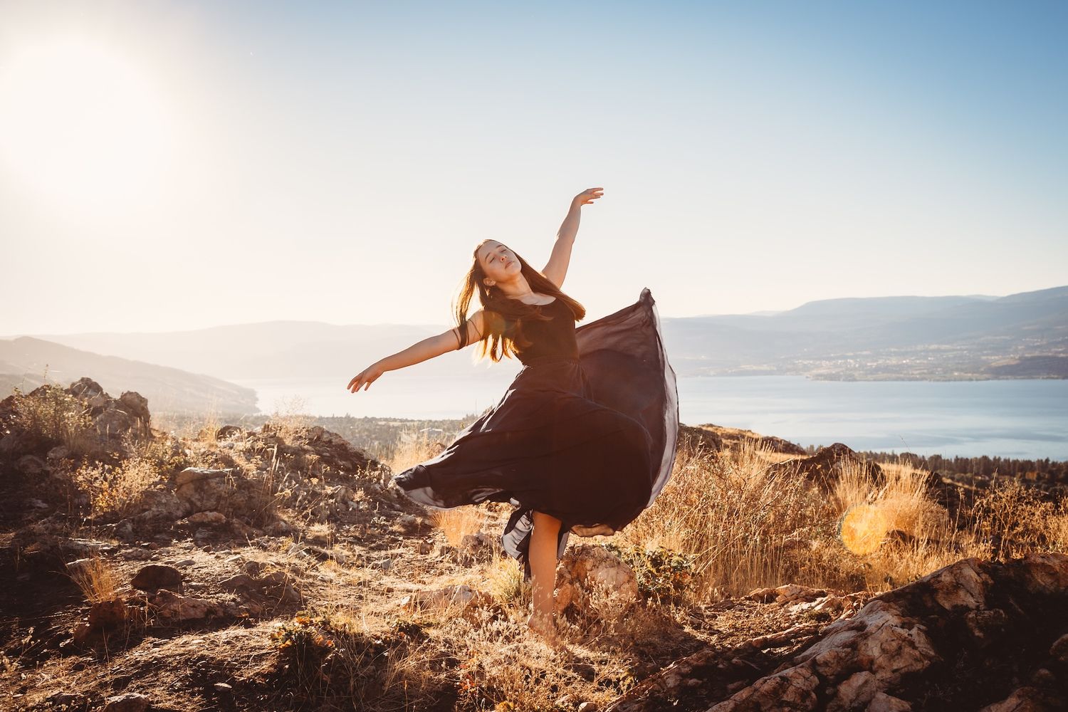 A dancer twirls in a flowing black dress on a cliff overlooking a serene mountain lake.
