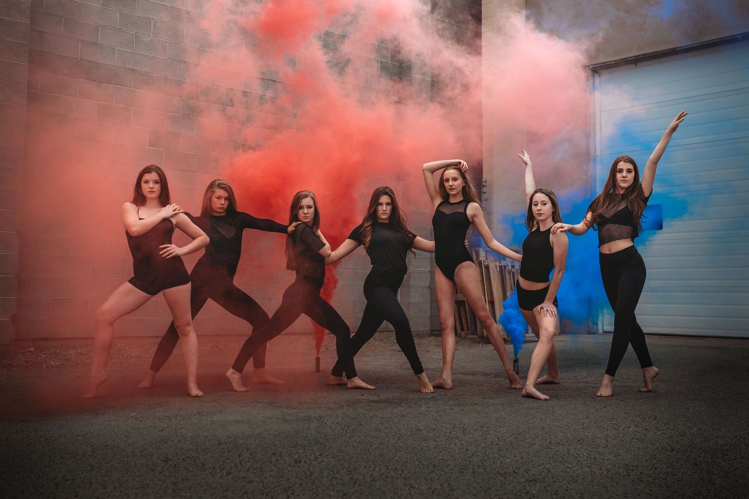 Dancers pose together with colorful smoke bombs creating vibrant pink and blue effects against an urban wall.