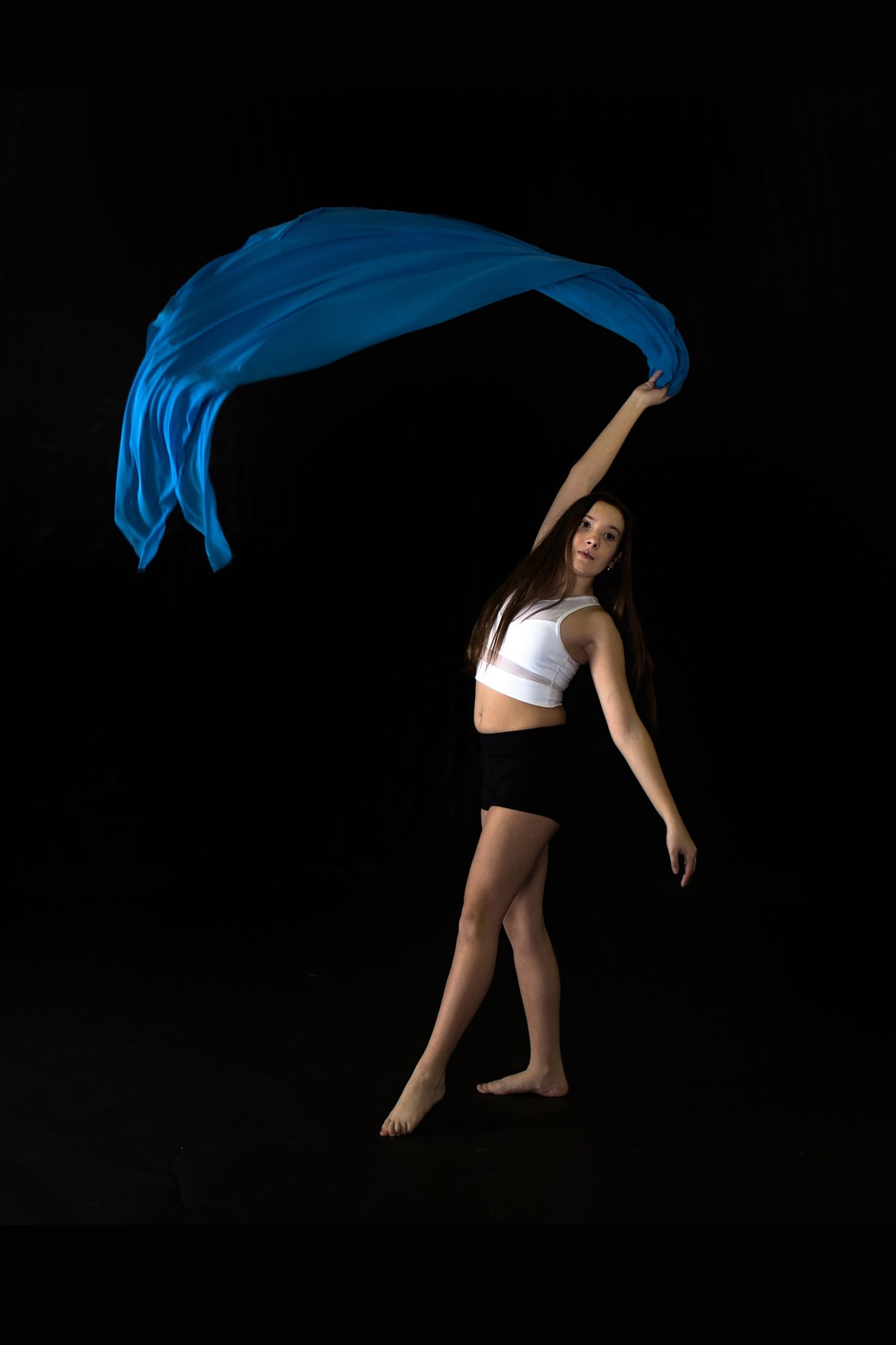 Dancer in white top and black shorts gracefully waves flowing blue fabric against dark background in artistic pose.