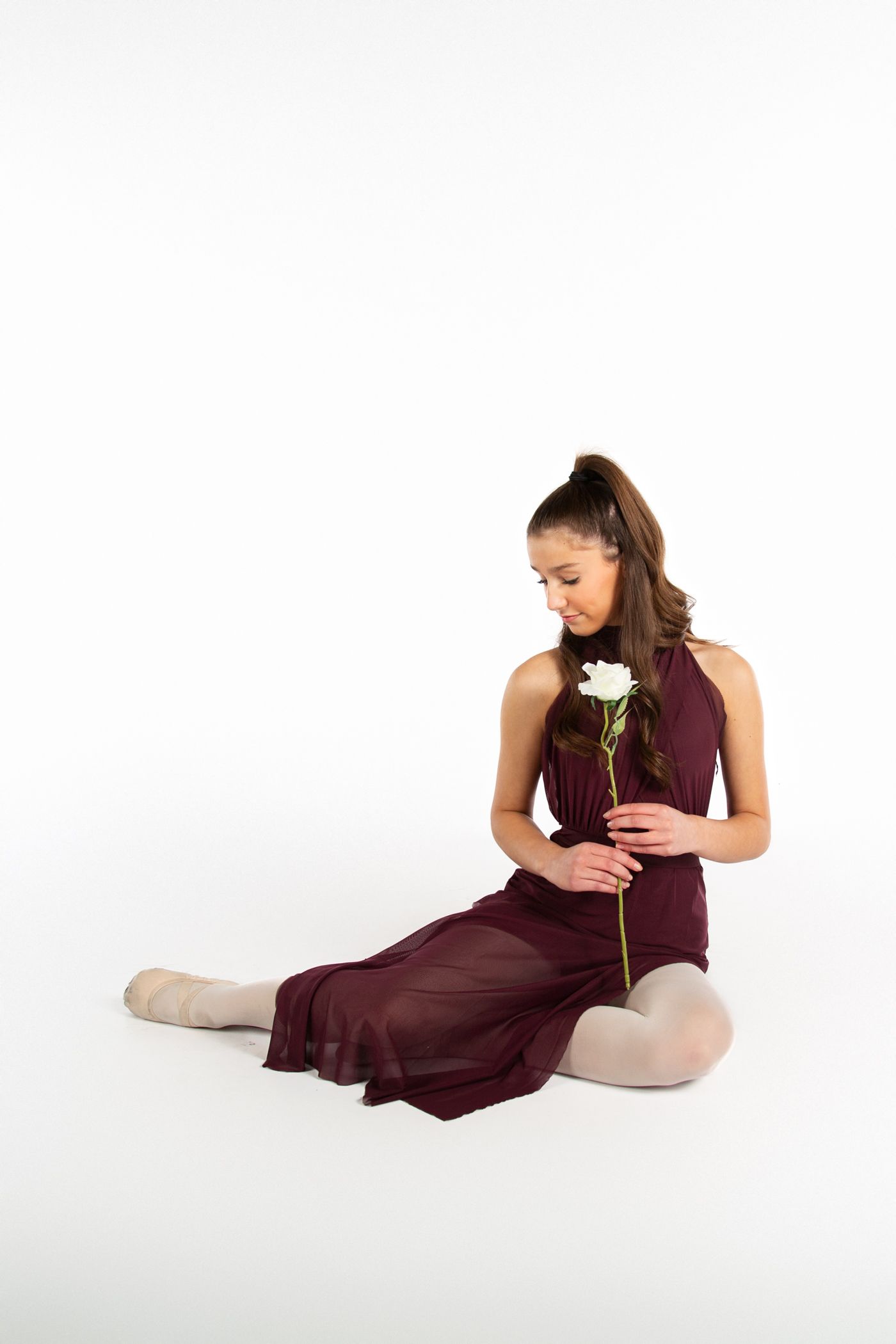 A dancer in a burgundy dress sits gracefully on the floor holding a single white rose while looking down thoughtfully.