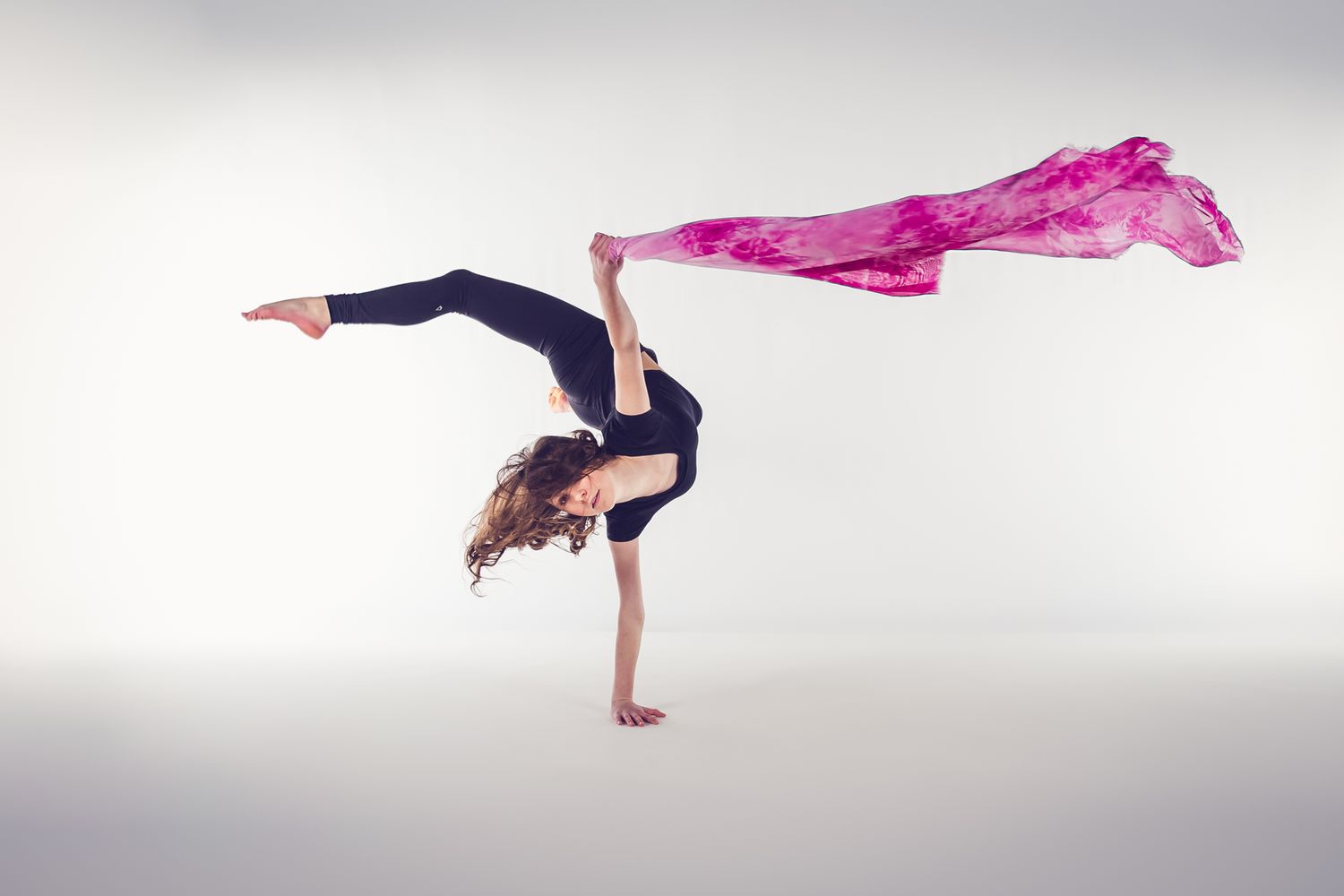 Dance performer in black leotard executes graceful handstand with flowing pink fabric trailing behind.