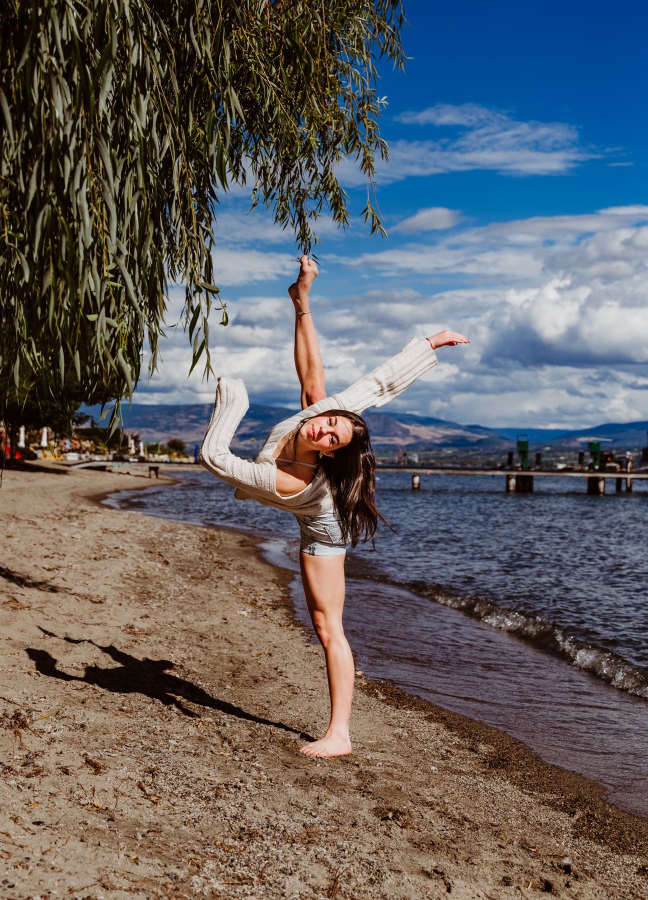 A dancer performs a dramatic standing split pose on a beach shoreline with willow trees and mountains in the background.