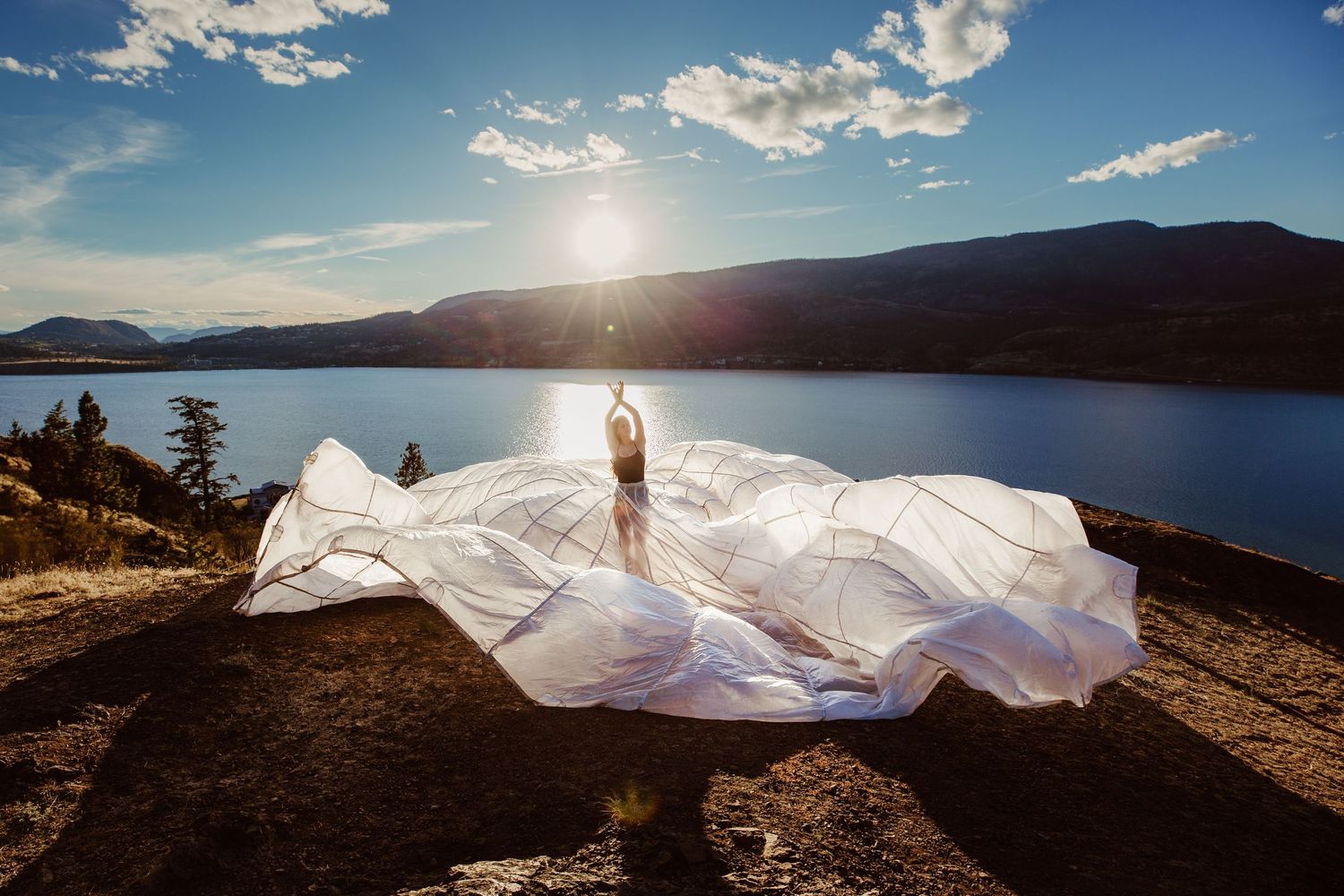 Flowing white wedding dress billows dramatically on cliff overlooking serene mountain lake at sunset.