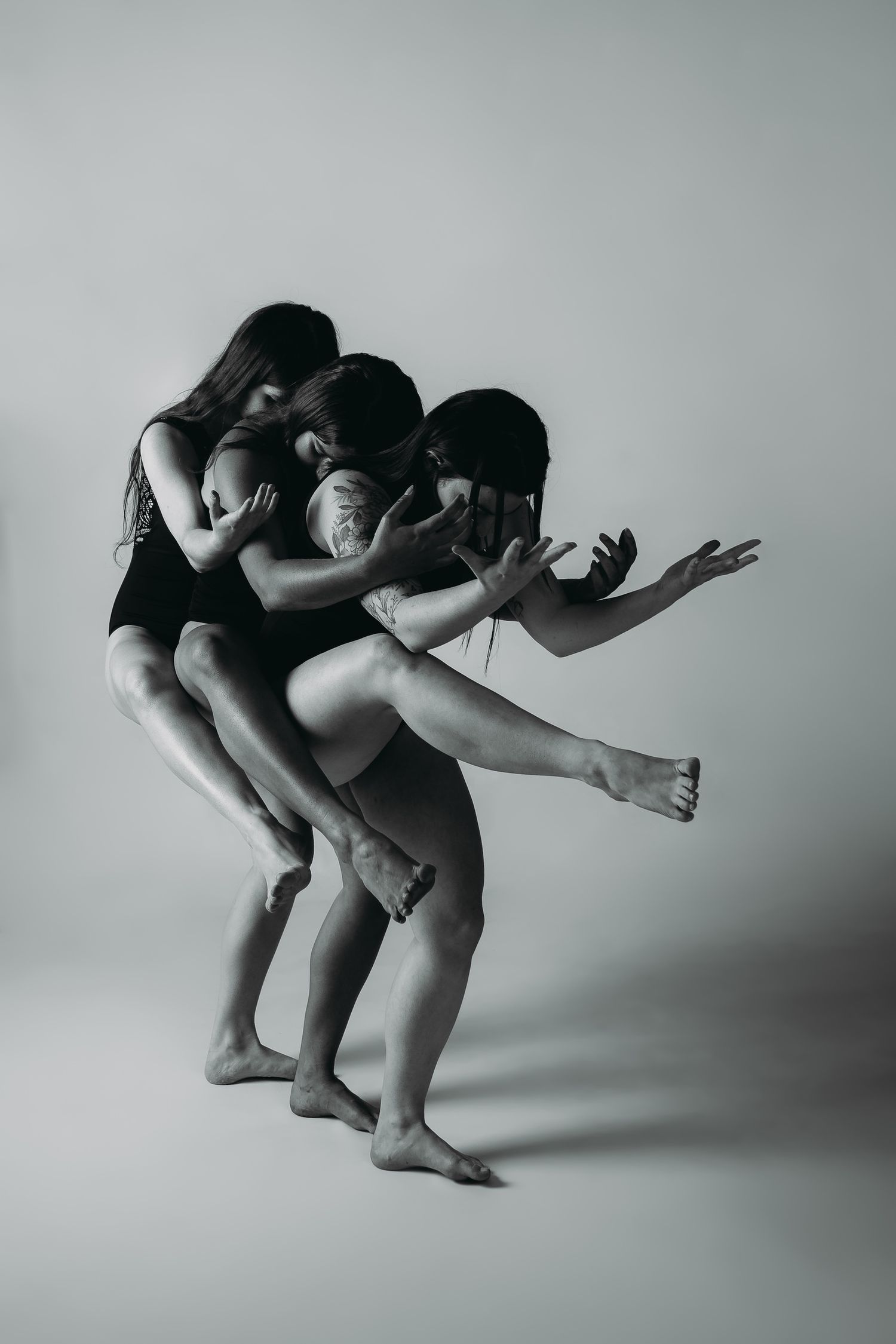 Three dancers in black leotards perform a contemporary dance movement against a gray background in dramatic lighting.