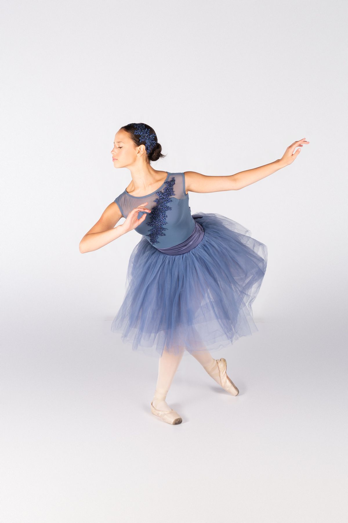 Ballet dancer in a flowing blue tulle dress performs a graceful pose against a white background.