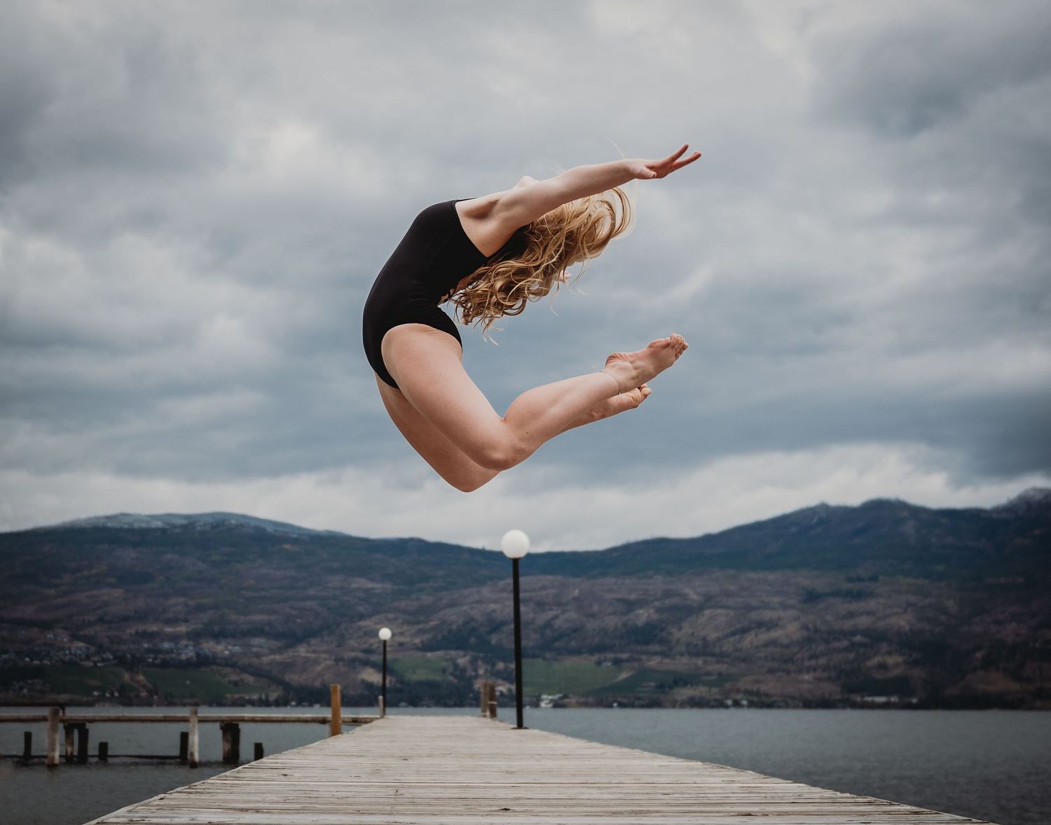 A dancer in a black leotard performs a graceful leap on a dock overlooking a mountainous lake on a cloudy day.