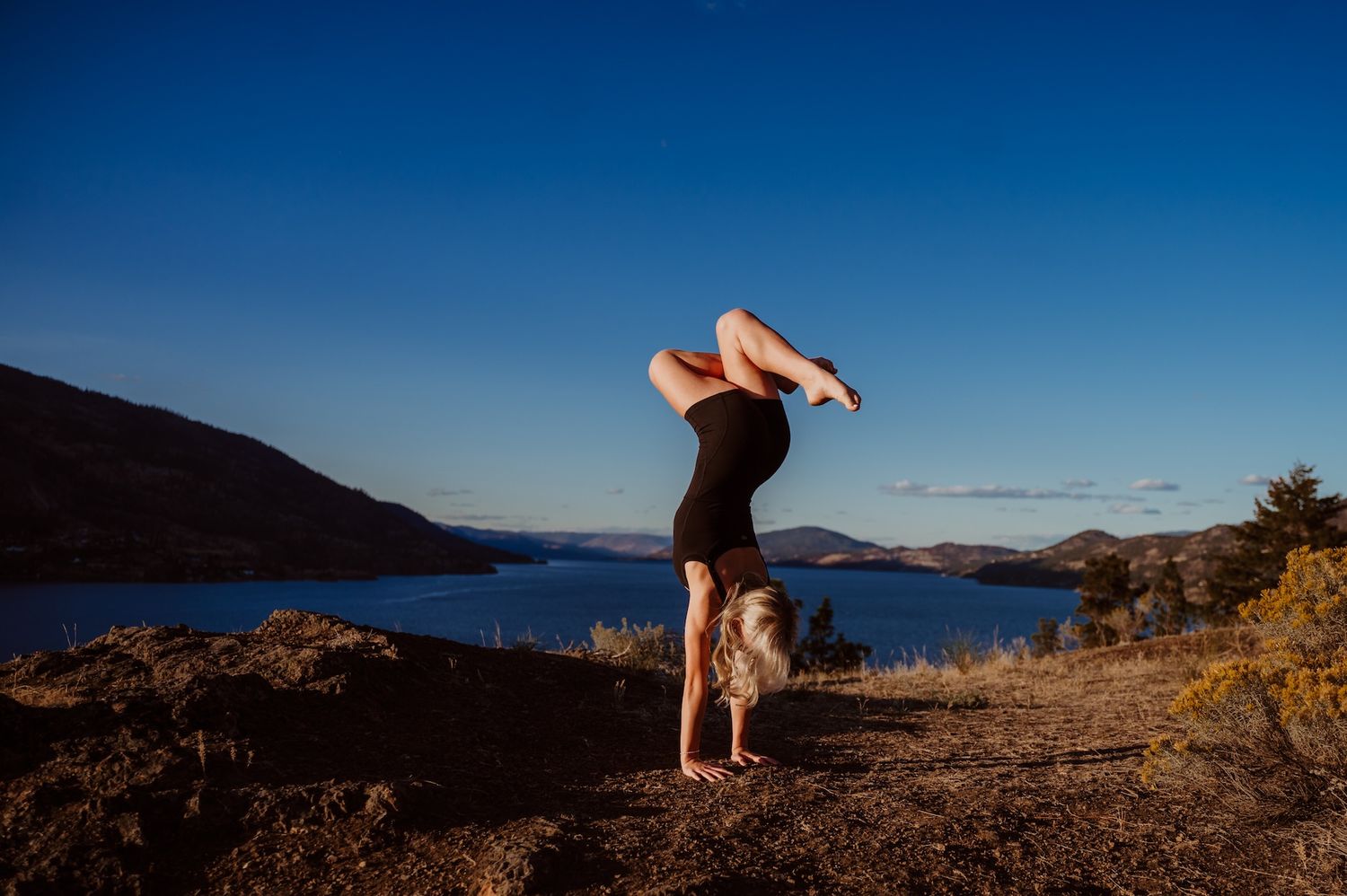 A person performs a handstand on rocky terrain overlooking a serene lake with mountains in the background at sunset.