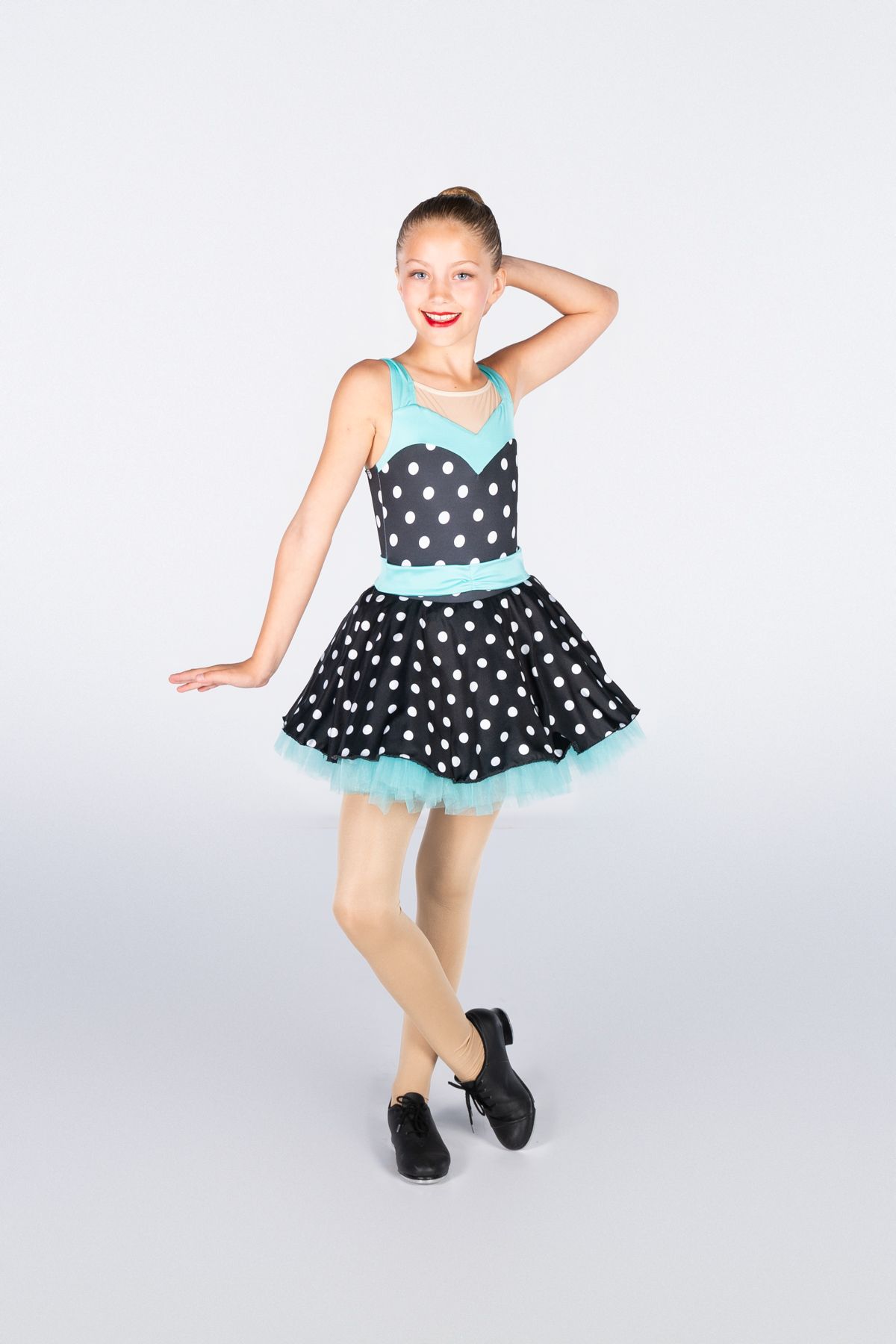 Dancer in polka dot dress with turquoise trim strikes playful pose against white backdrop.
