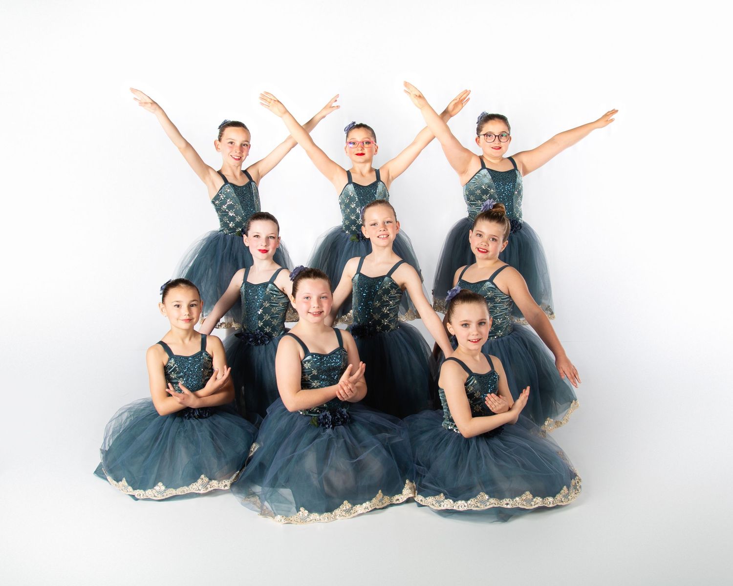 Ballet dancers in dark teal tutus pose gracefully in a group formation against a white backdrop.