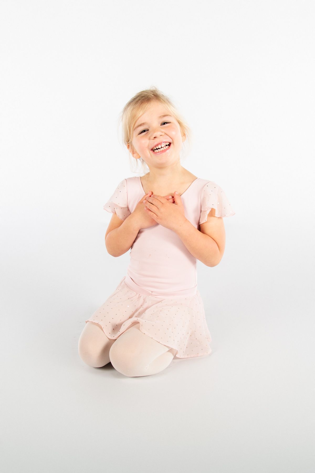 A joyful young dancer in a pink leotard kneeling and smiling against a white background.