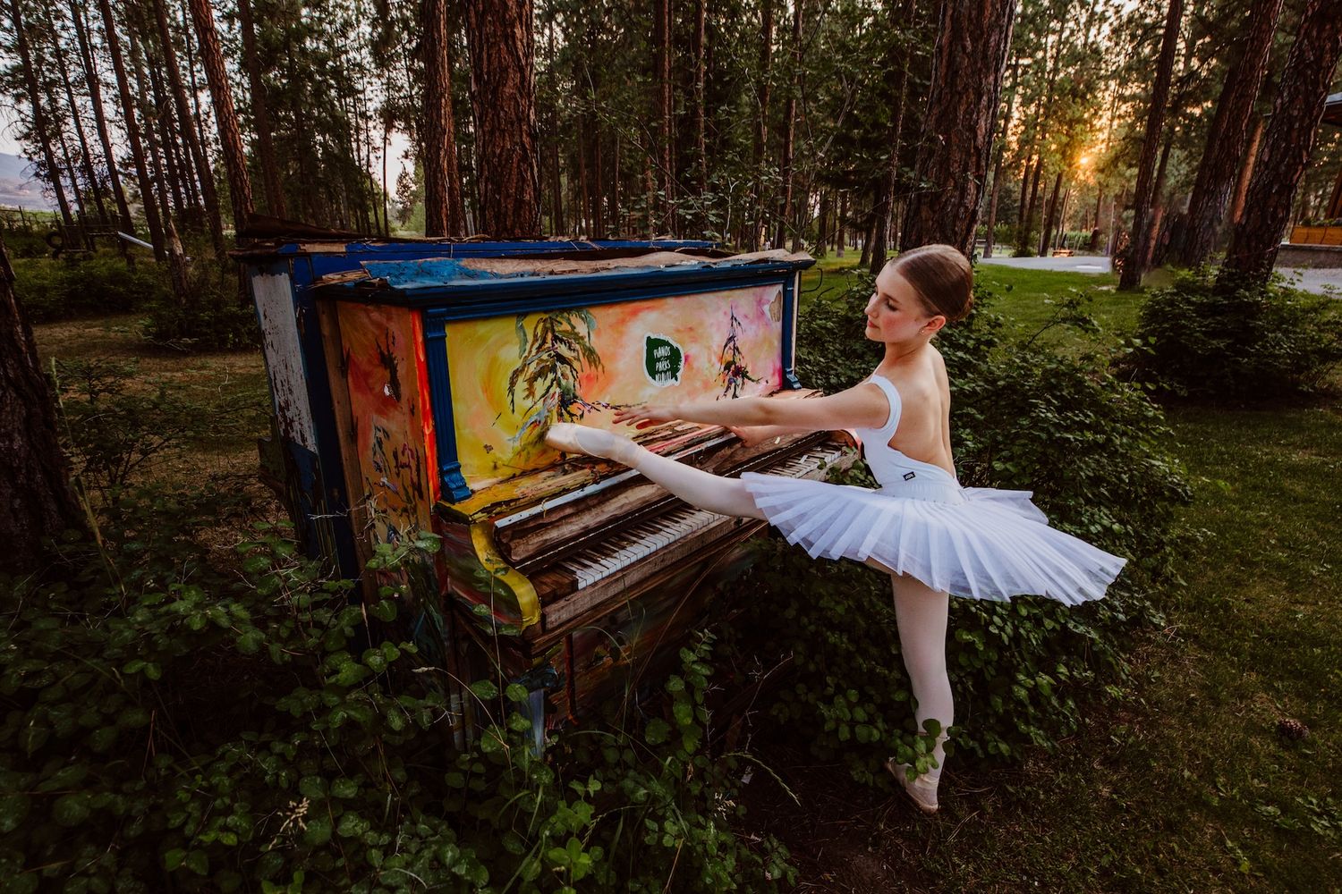 A ballerina in a white tutu poses gracefully next to a colorful graffiti-covered piano in a sunlit forest setting.
