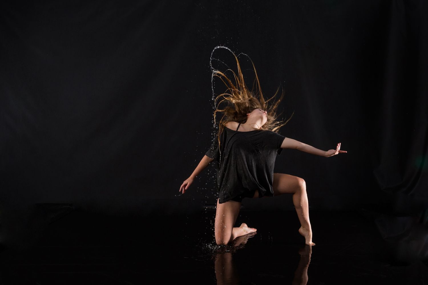 A dancer in black dress performs a dynamic leap with hair in motion against a dark background.