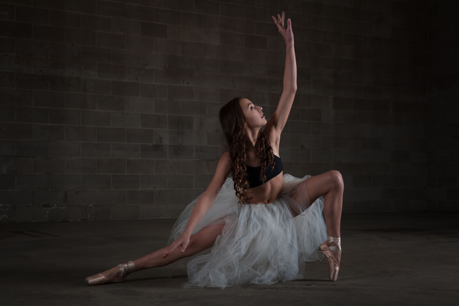 Ballet dancer in a white tulle dress performs an elegant floor pose against a dark brick wall.