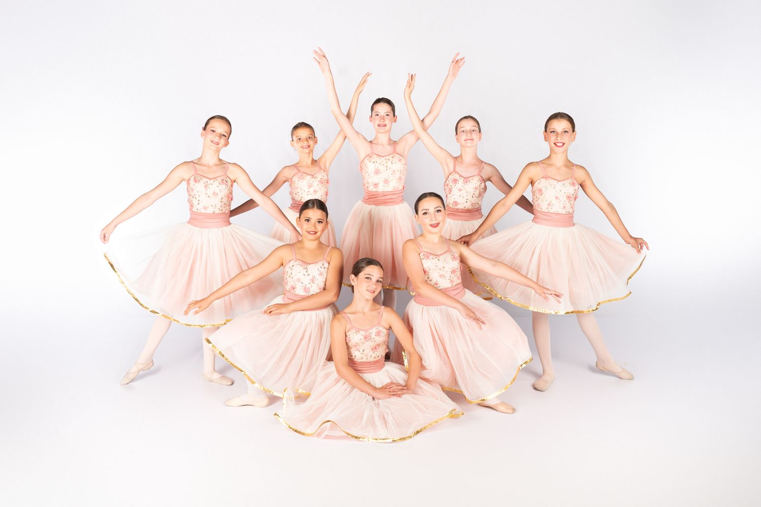 Ballet dancers in peach-colored tutus pose gracefully in a group formation against a light backdrop.