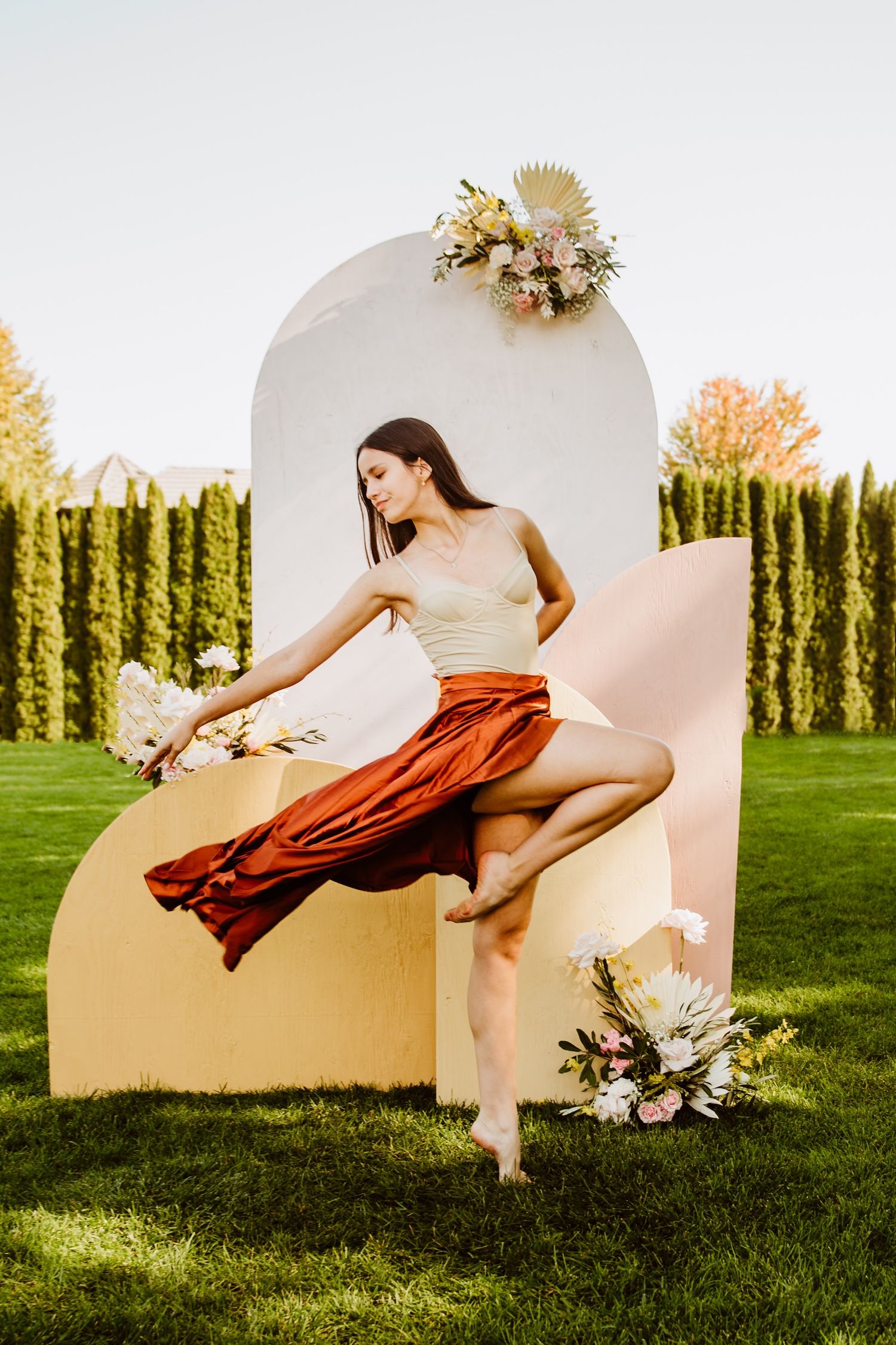 Dancer in flowing red skirt leaps gracefully against curved backdrop with dried floral arrangements.
