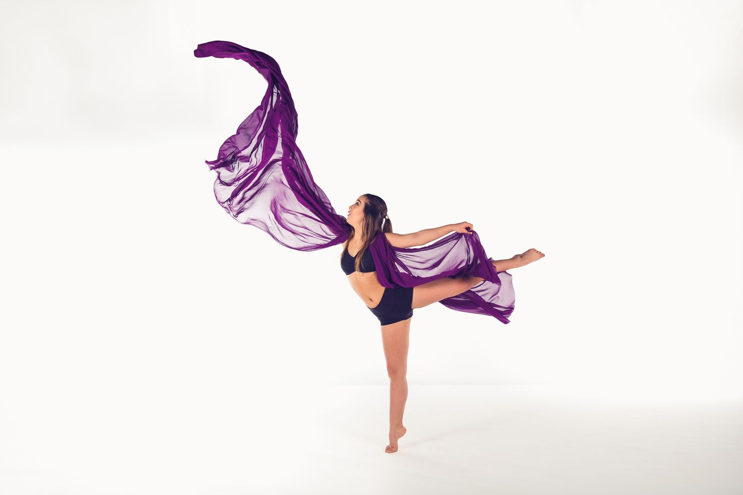 Dancer strikes graceful pose in black outfit with flowing purple fabric creating dramatic movement against white background.
