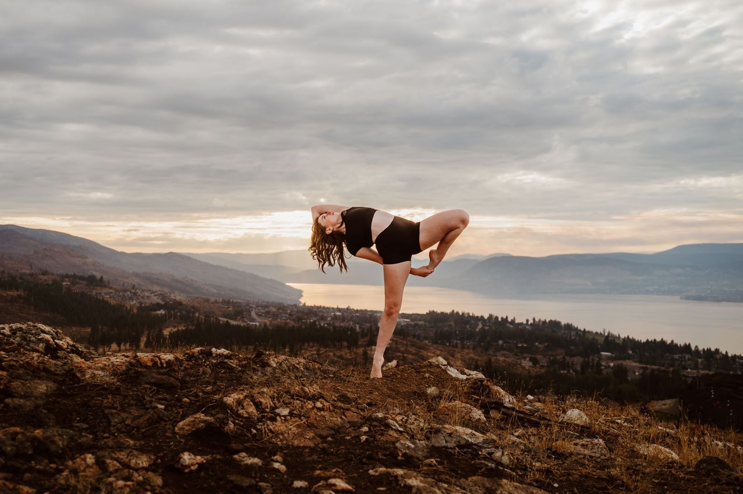 A person practices yoga in a half moon pose on a mountain ridge overlooking a misty lake at sunset.