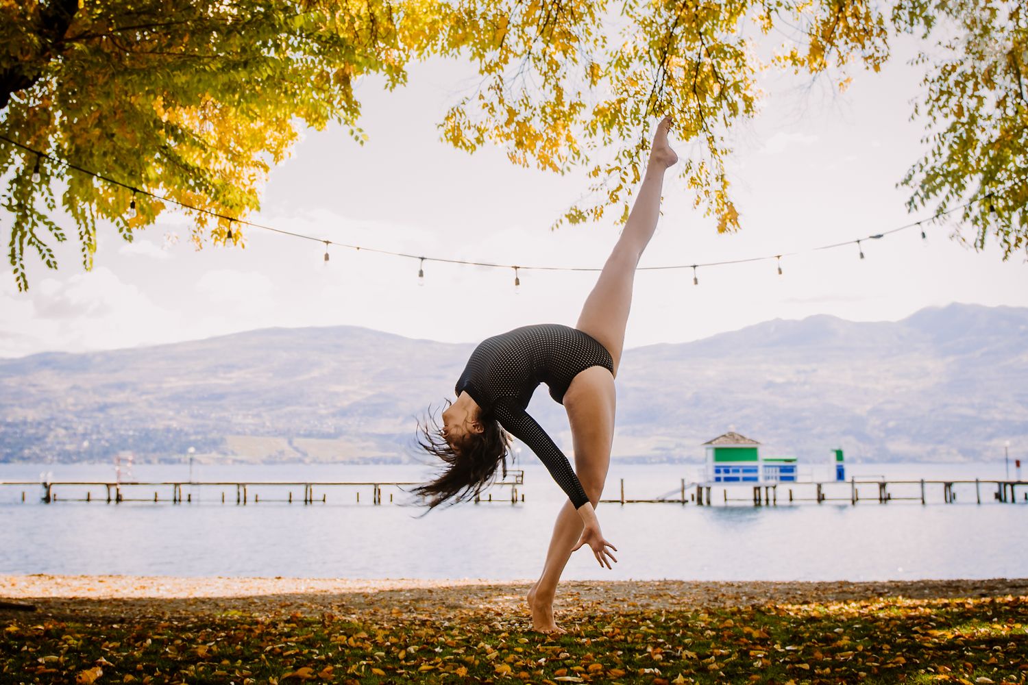 A dancer performs a graceful backbend on a lakeside path during autumn with mountains and string lights visible in background.