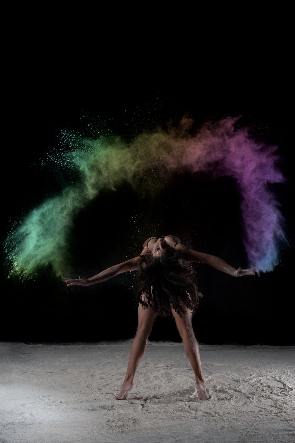 Dancer performs dramatic pose amid colorful powder explosion of green and purple against black background.