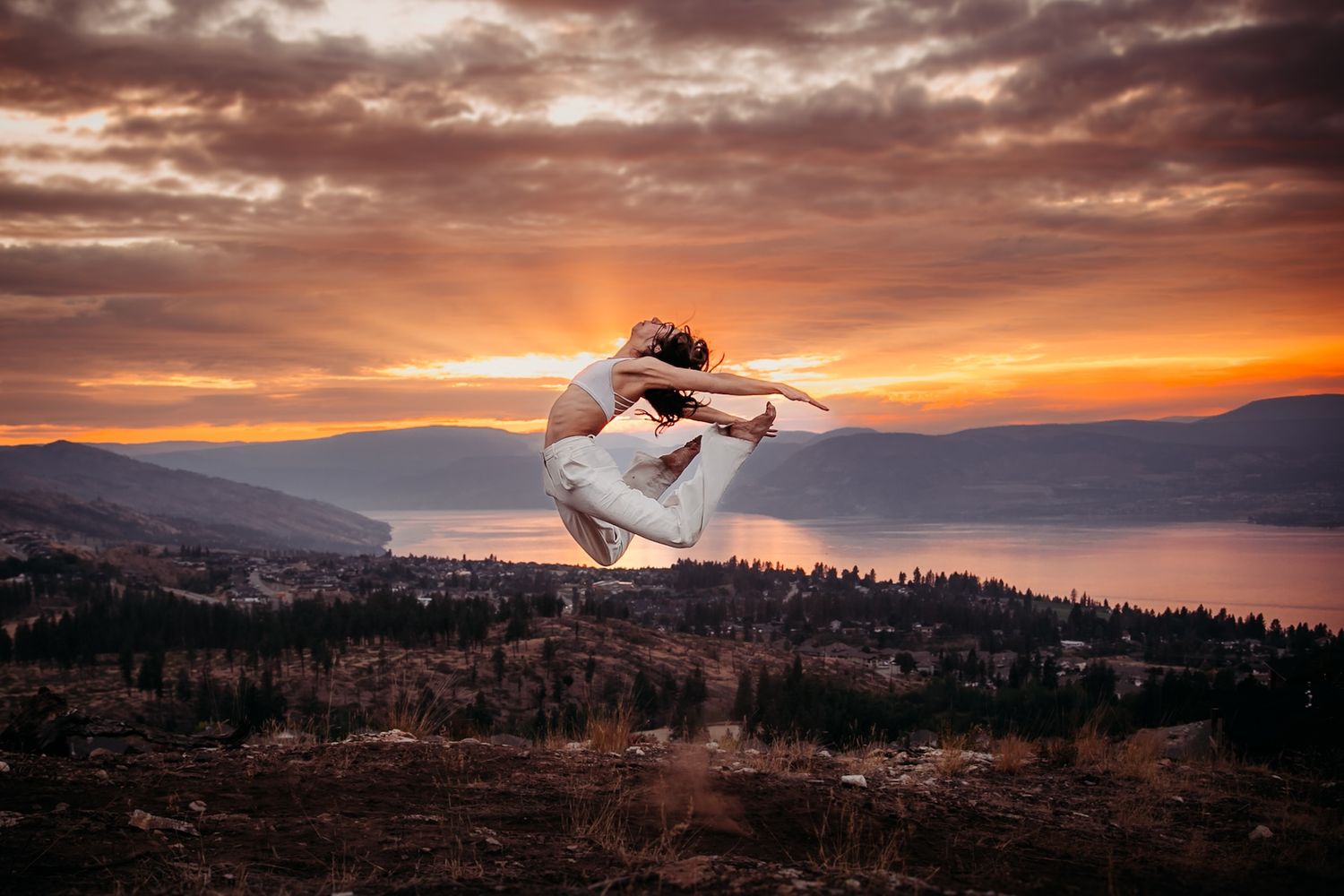 A dancer leaps gracefully against a dramatic sunset over a mountainous lake landscape.