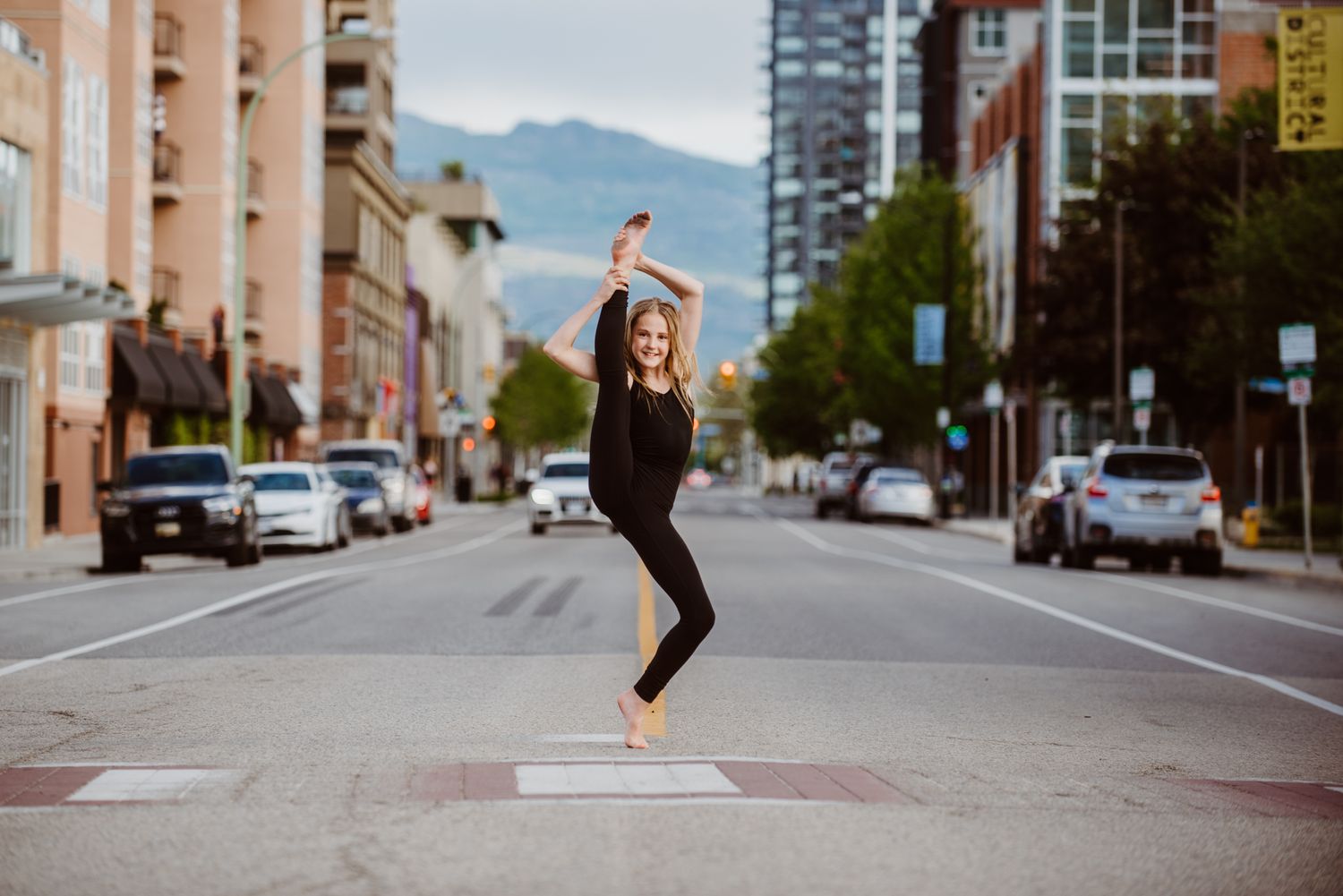 A dancer in black athletic wear performs a graceful leap on a city street with mountains visible in the background.