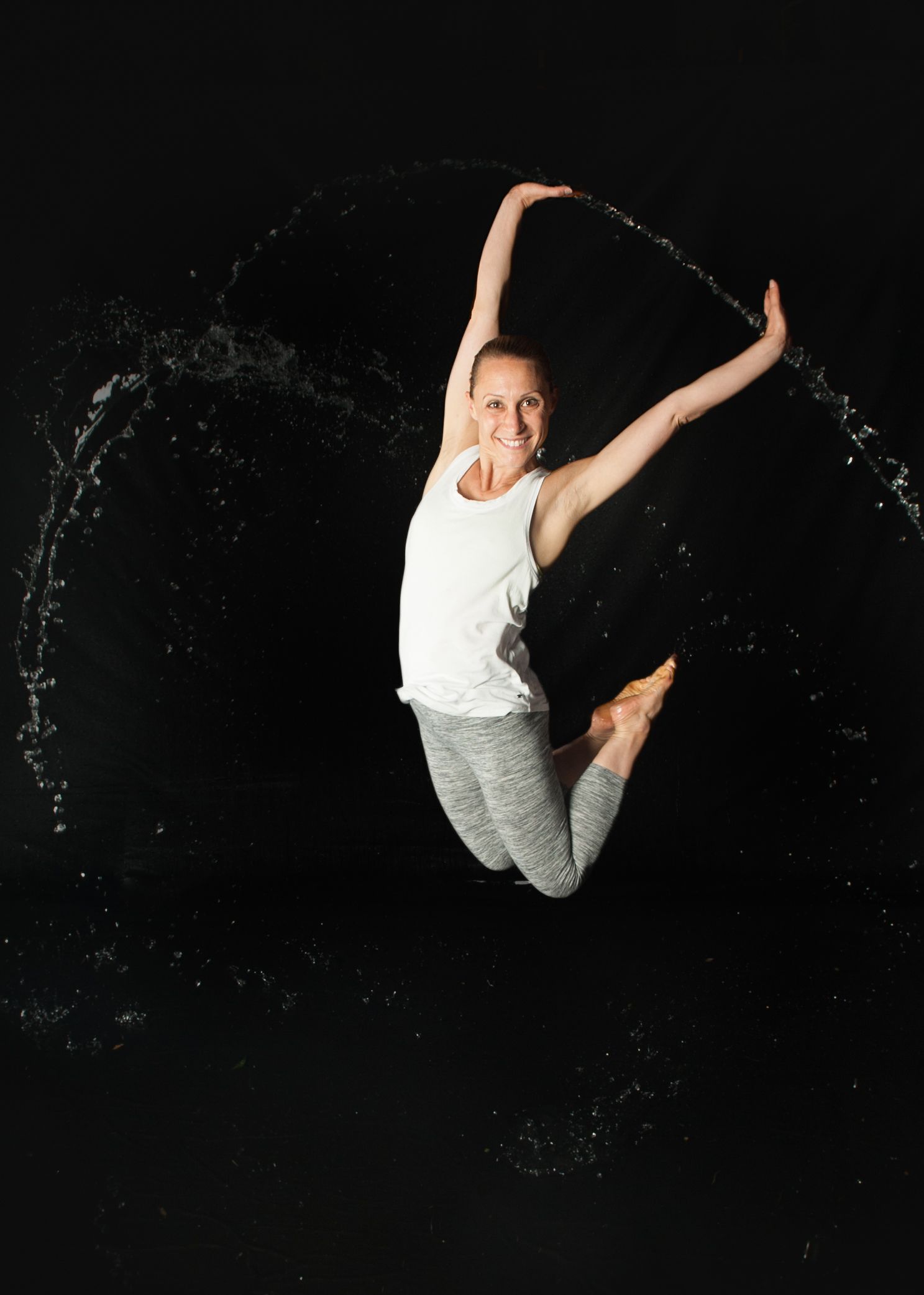 Dancer leaping mid-air wearing white top and gray pants with water arcs against black backdrop.