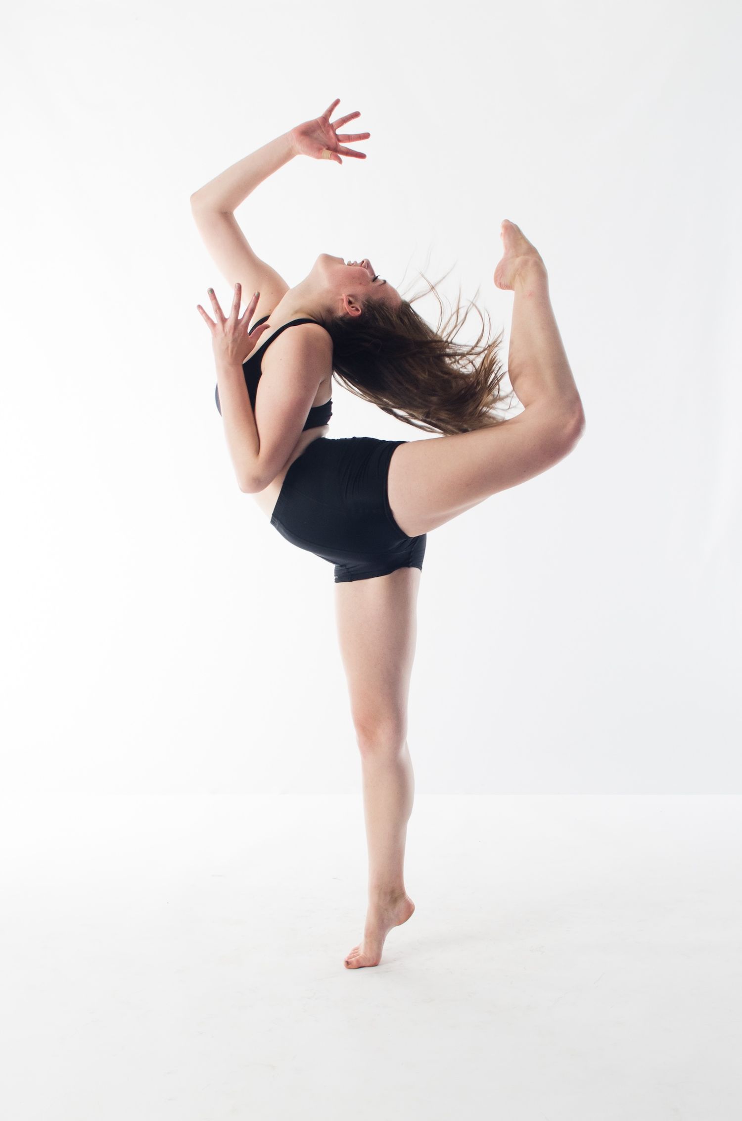 Contemporary dancer in black leotard performs a dynamic high kick with expressive arm movements against white backdrop.