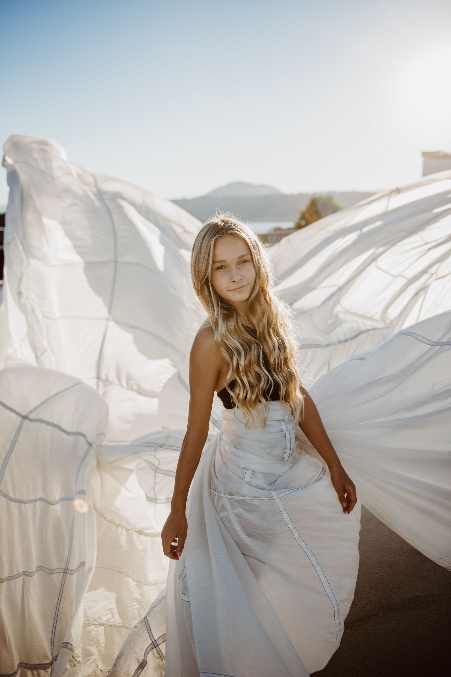 Young woman in flowing white dress stands with windswept fabric billowing behind her against sunny sky.