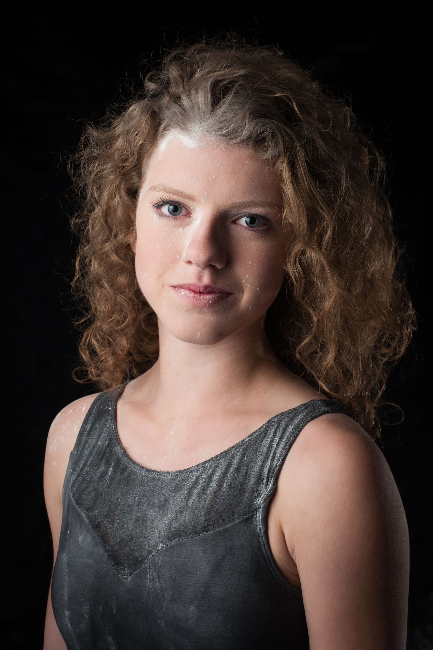 Portrait against black background showing curly hair and grey tank top in dramatic lighting.