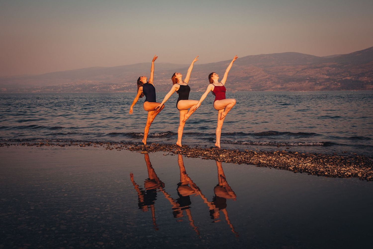 Three ballet dancers perform arabesques in sunset light on a beach with their reflections mirrored in the wet sand.